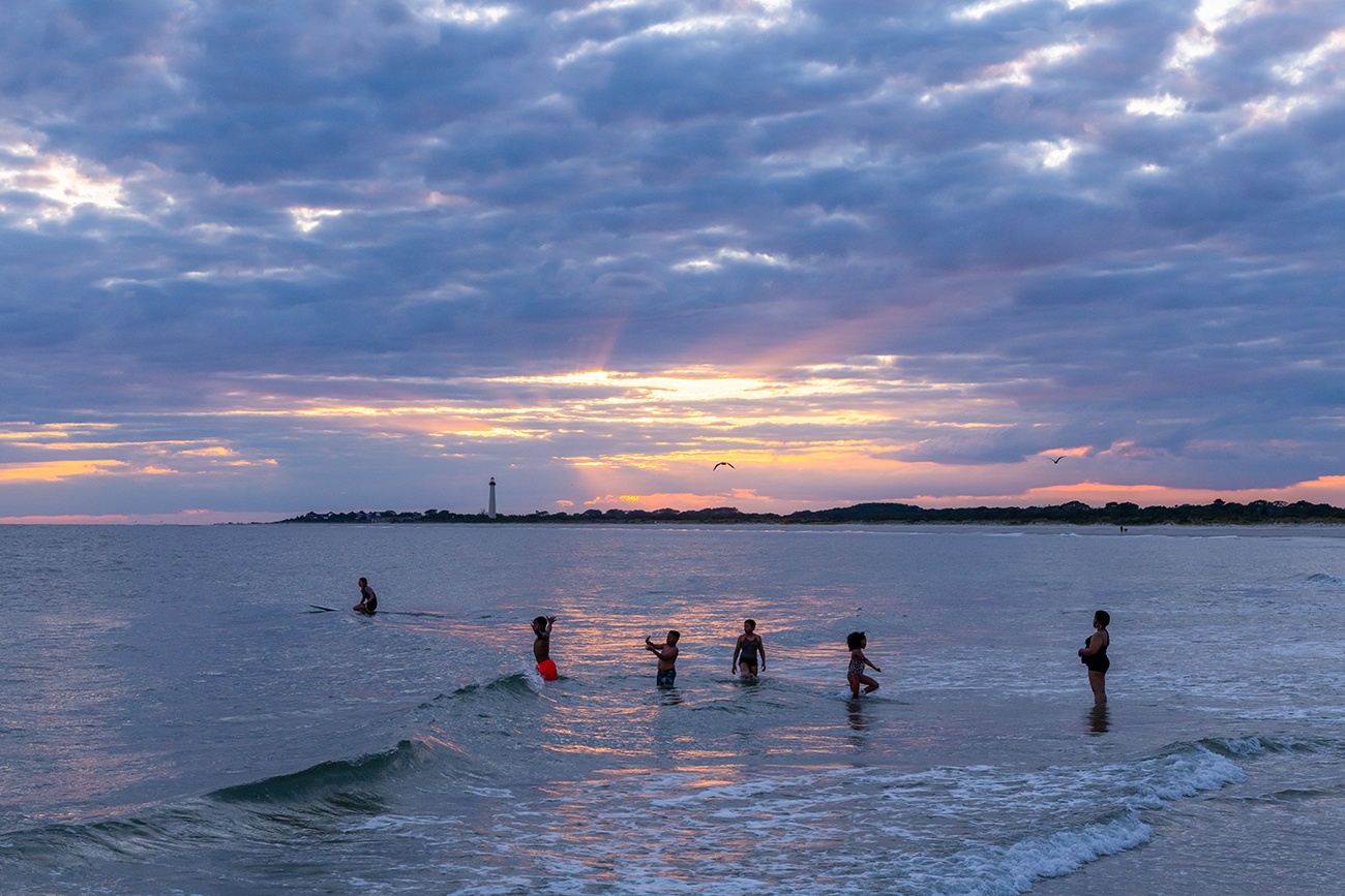 Kids playing in the ocean and a person on a surfboard as the sun sets behind clouds with the Cape May lighthouse in the distance