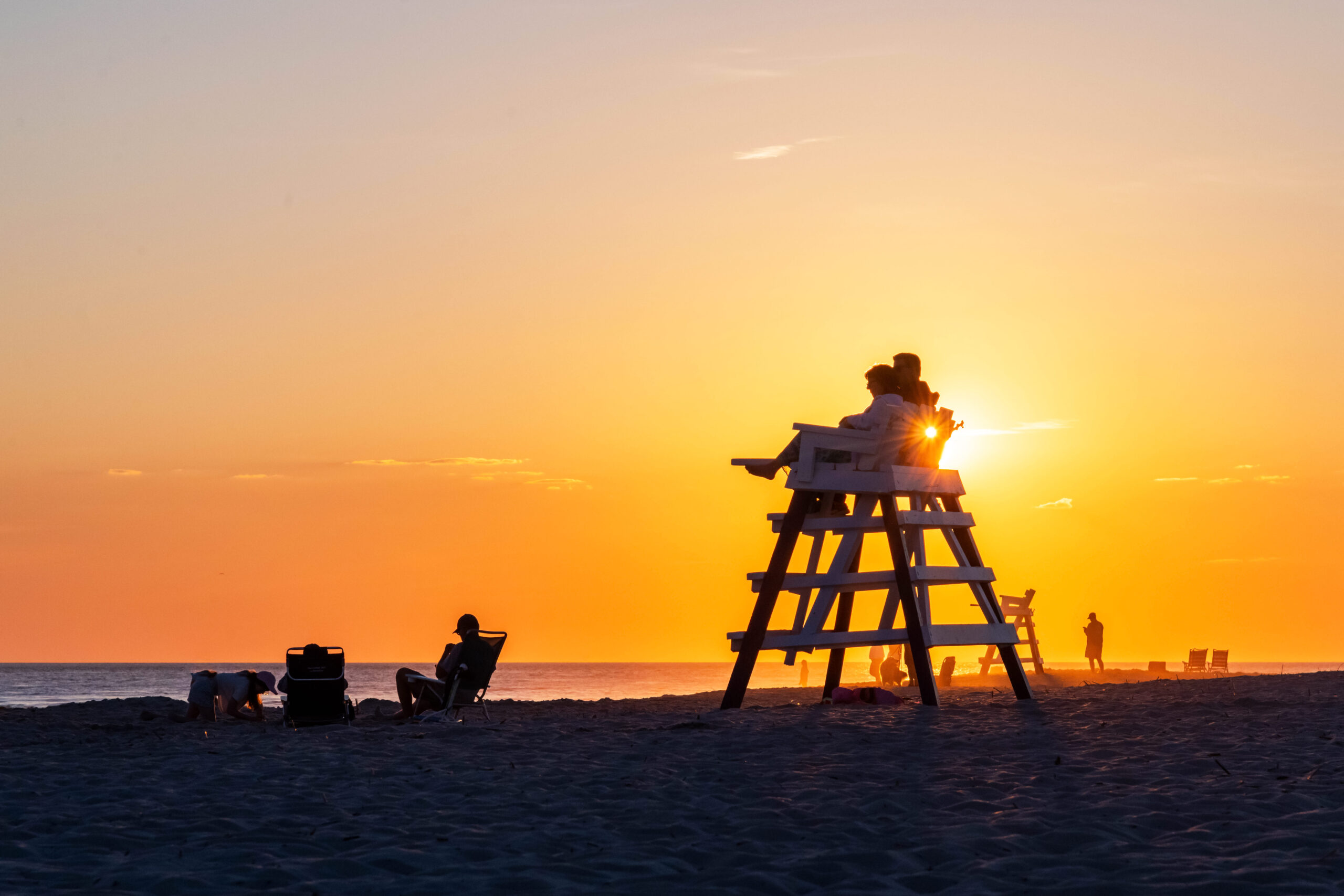 The sun setting directly behind two people sitting on a lifeguard stand. Other groups are sitting on the beach, and people are watching sunset in the distance