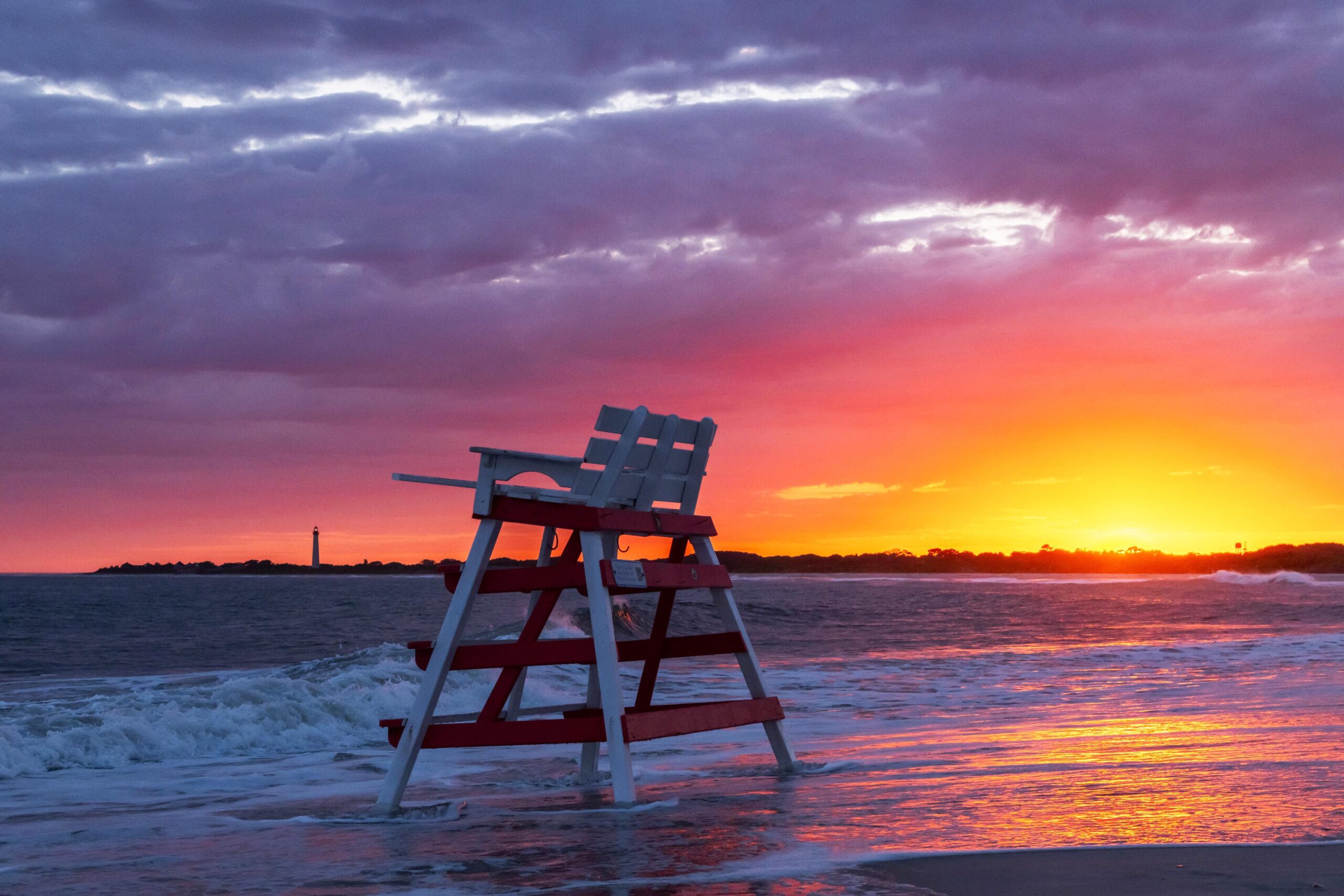 The ocean rushing into the beach at sunset with brilliant yellow, orange, pink, and purple clouds in the sky. The lifeguard stand is at the ocean's edge, and the Cape May lighthouse is in the distance.