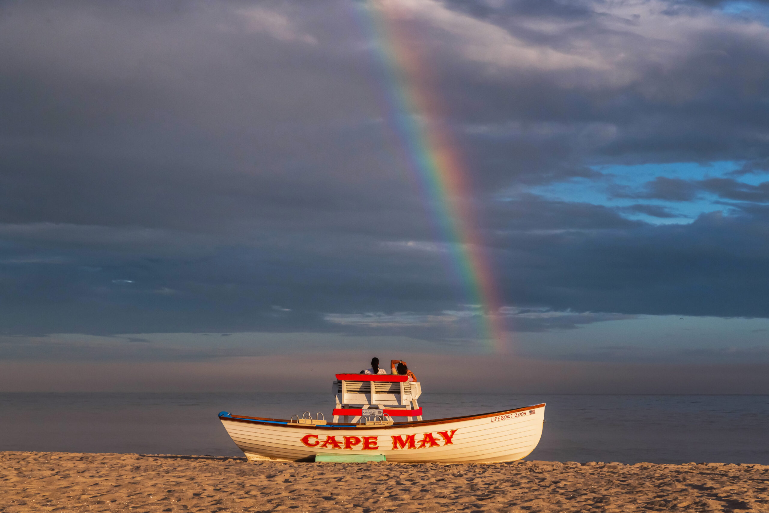 A rainbow is in a cloudy sky. There are two people sitting in a lifeguard stand and there is a "Cape May" lifeguard boat on the beach.