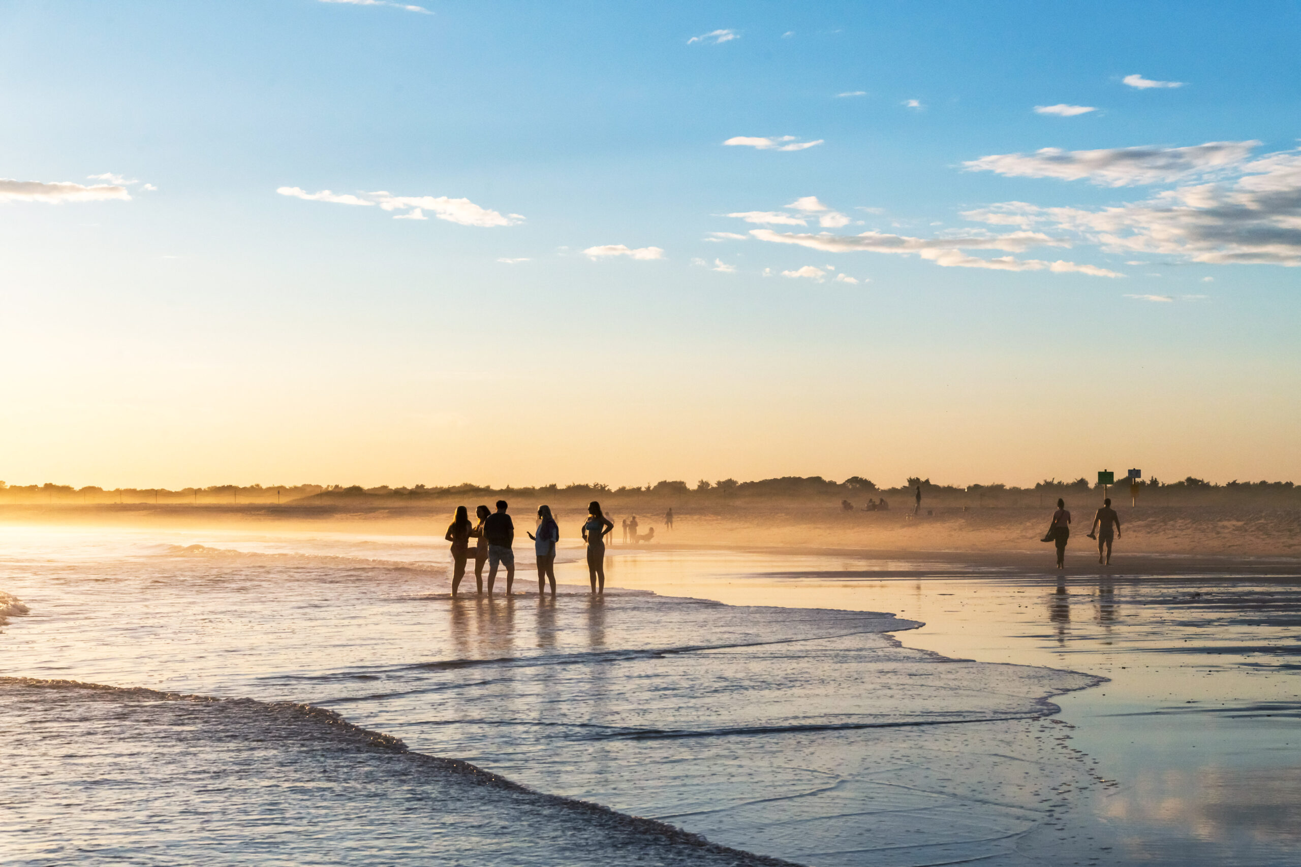 People standing at the water's edge of the ocean and walking on the beach. The sky is blue and gold and there is a slight mist coming off the ocean.