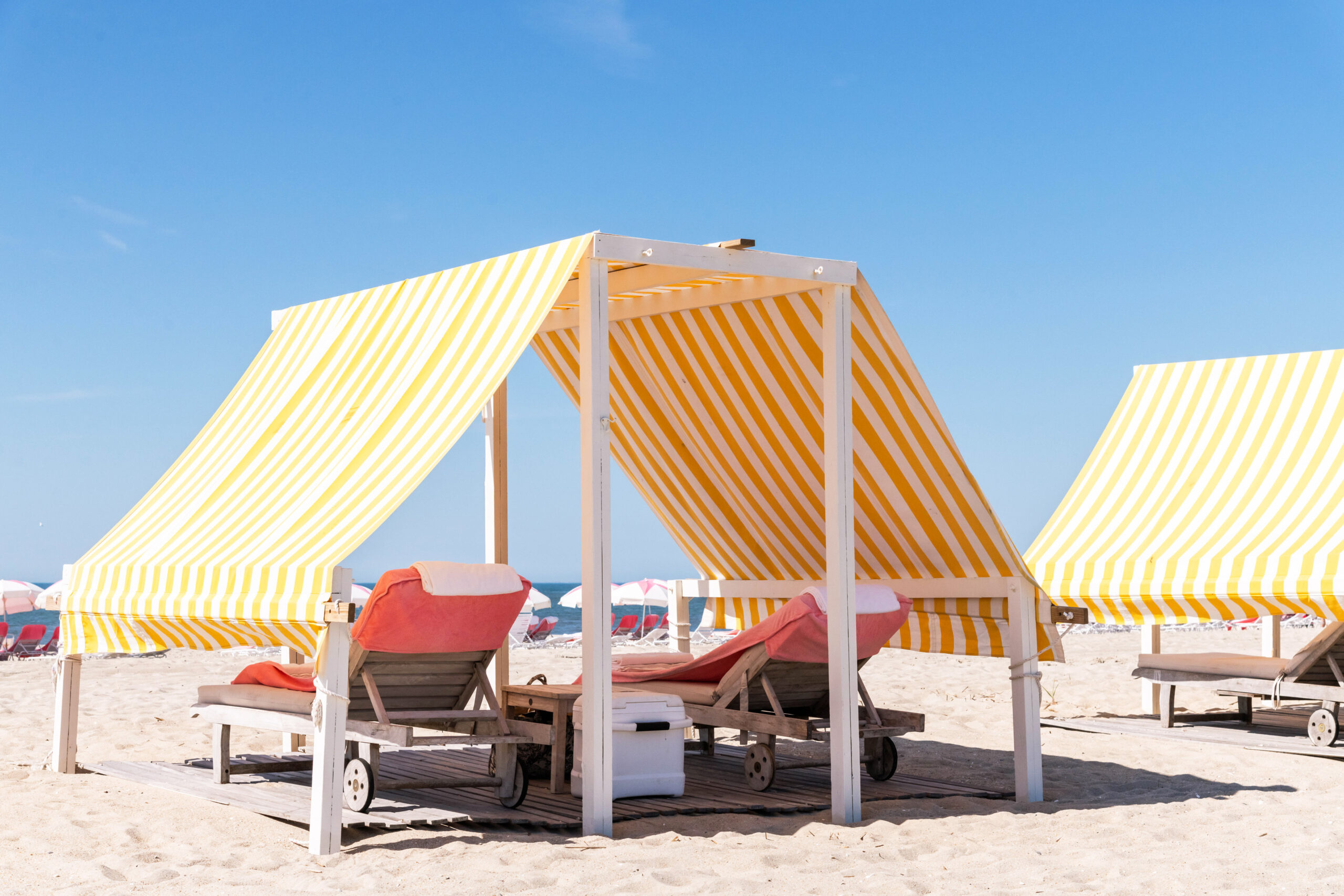 A yellow and white striped beach cabana covering two pink lounge chairs on the beach. It is a sunny bright day with a blue sky.