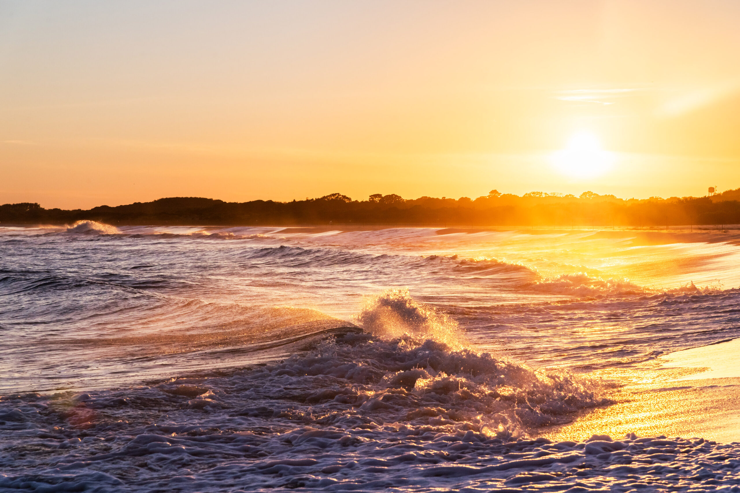 The sun setting and shining on a wave crashing into the sand.