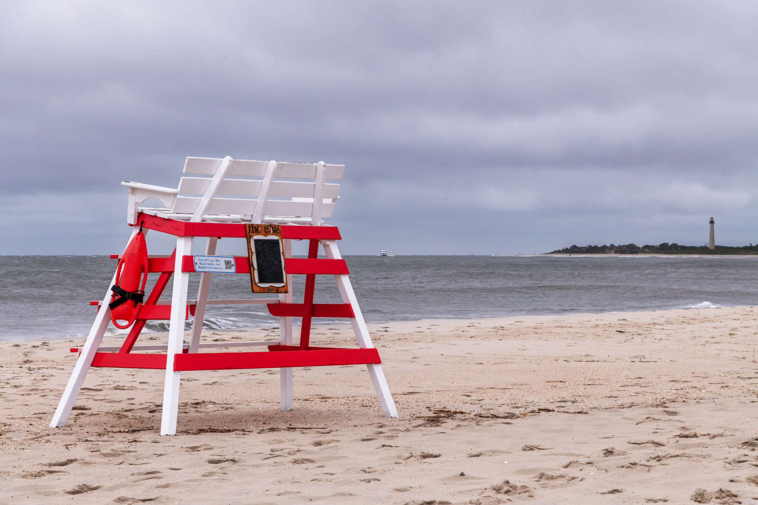 An empty lifeguard stand on the beach with clouds in the sky and the Cape May Lighthouse in the background.