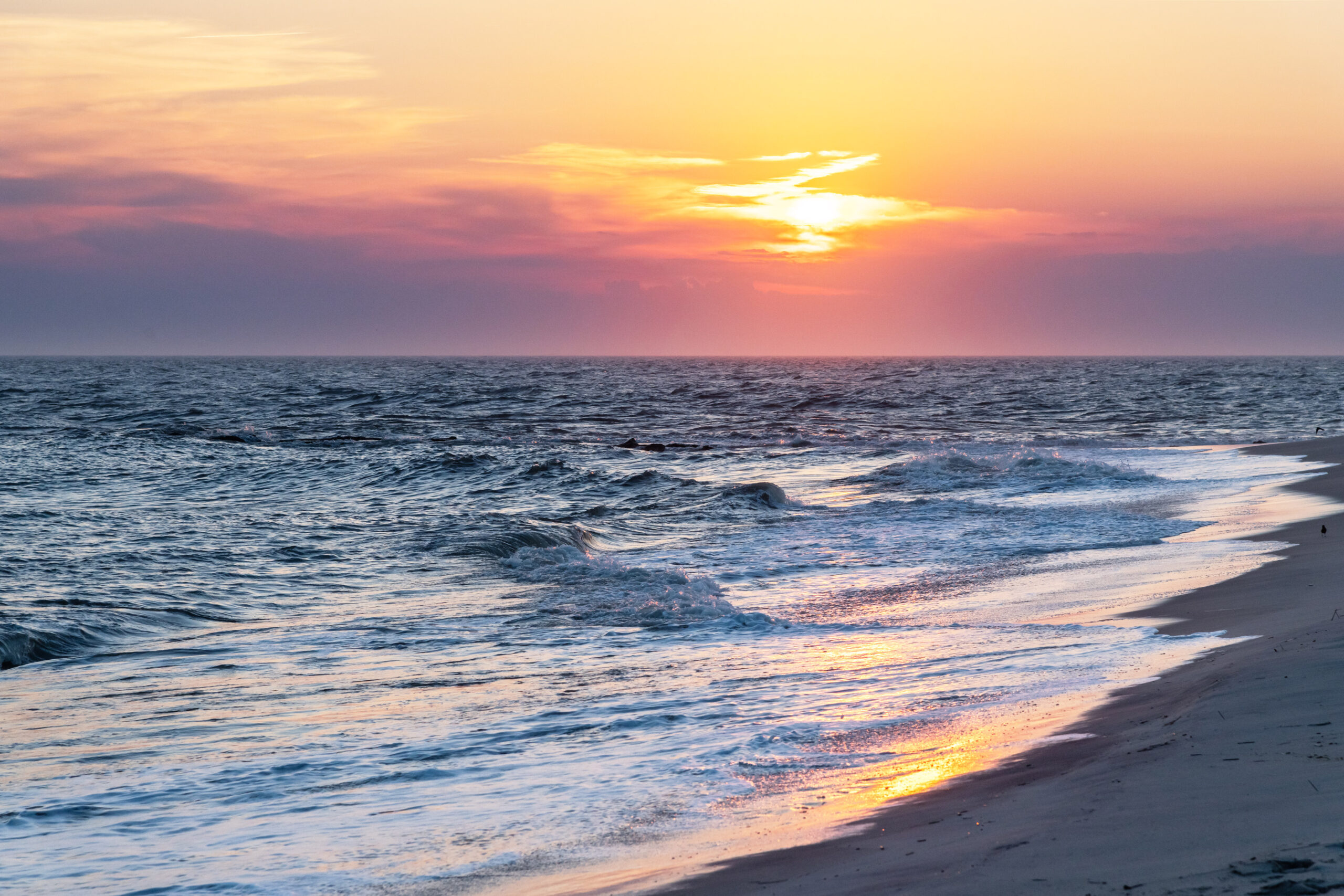 The sun setting behind hazy pink and purple clouds at the horizon line. The colors are reflected in the blue ocean crashing onto the shore.