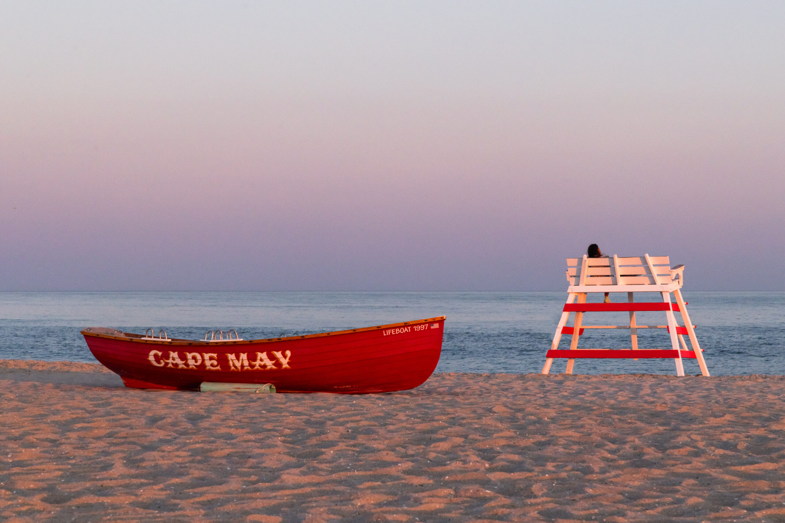 A red Cape May lifeguard boat and a Cape May lifeguard stand on the beach at sunset. Someone is sitting in the lifeguard stand, and the sky is clear, pink, and blue.