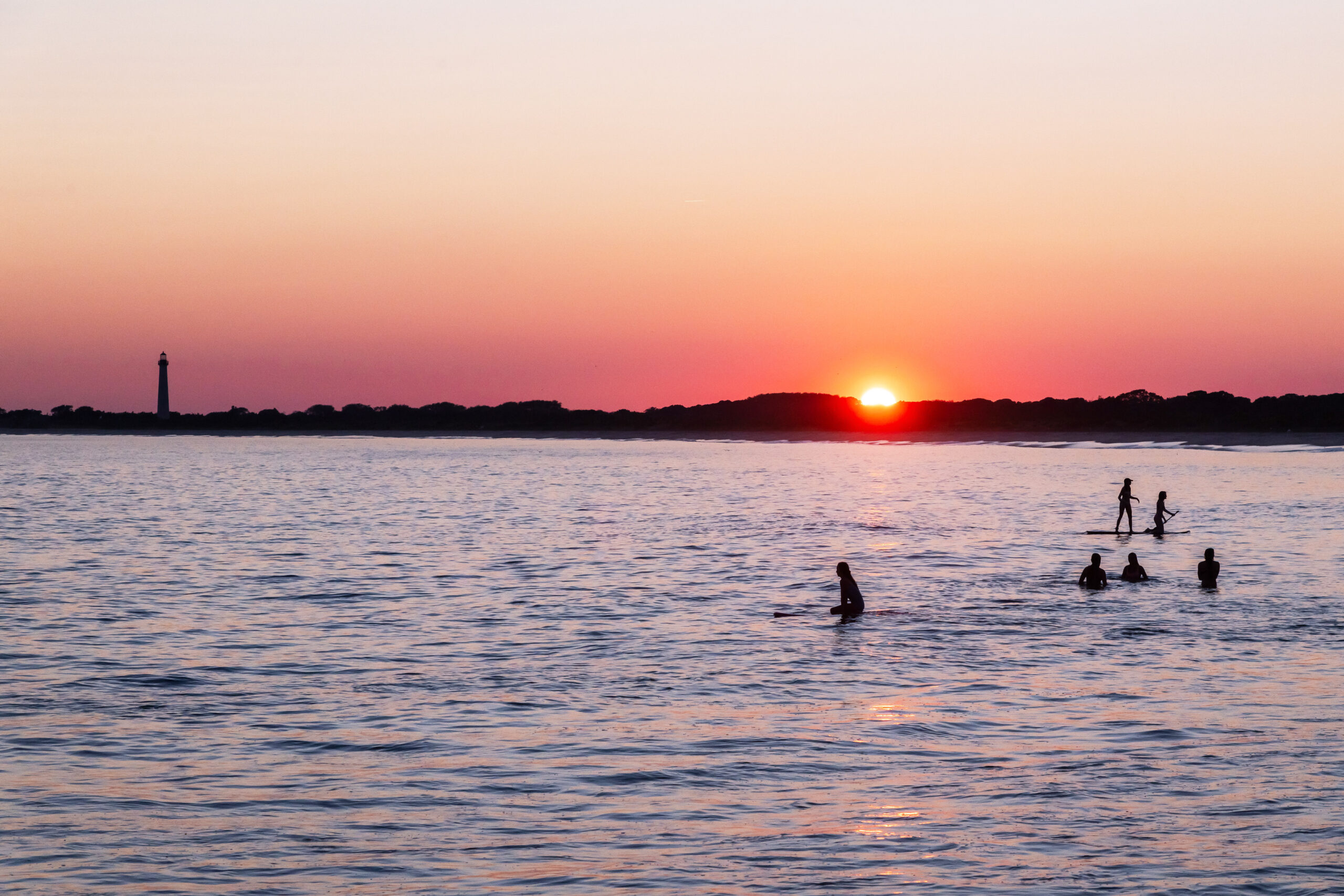 Silhouettes of people on surf boards and standing in the ocean as the sun sets with the Cape May Lighthouse. The sky is clear and pink.