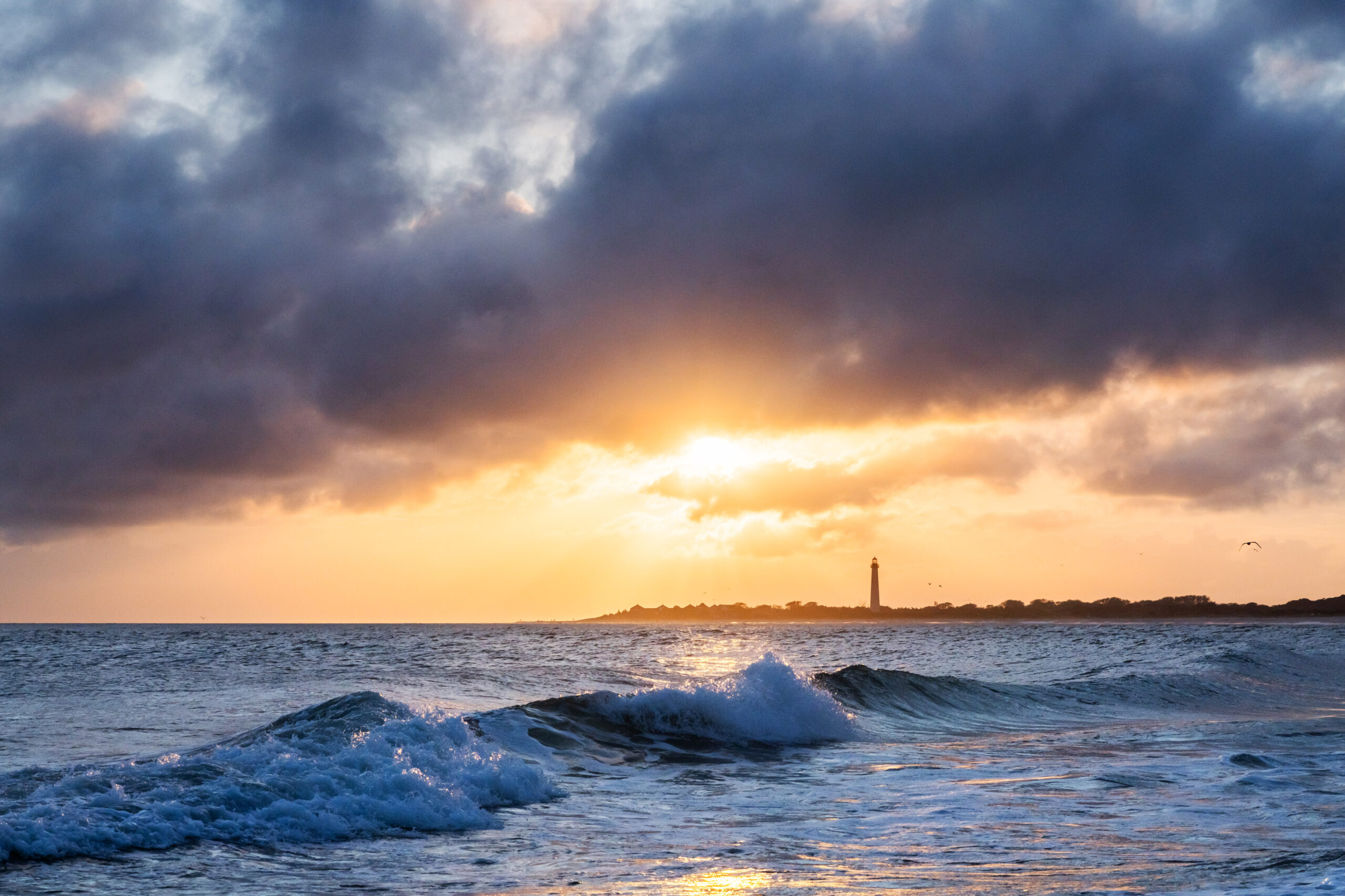 Puffy dark clouds in the sky with the sun setting peaking behind the clouds. The clouds are dark purple and the sun is golden. The Cape May Lighthouse is in the distance and a wave is crashing in the ocean.