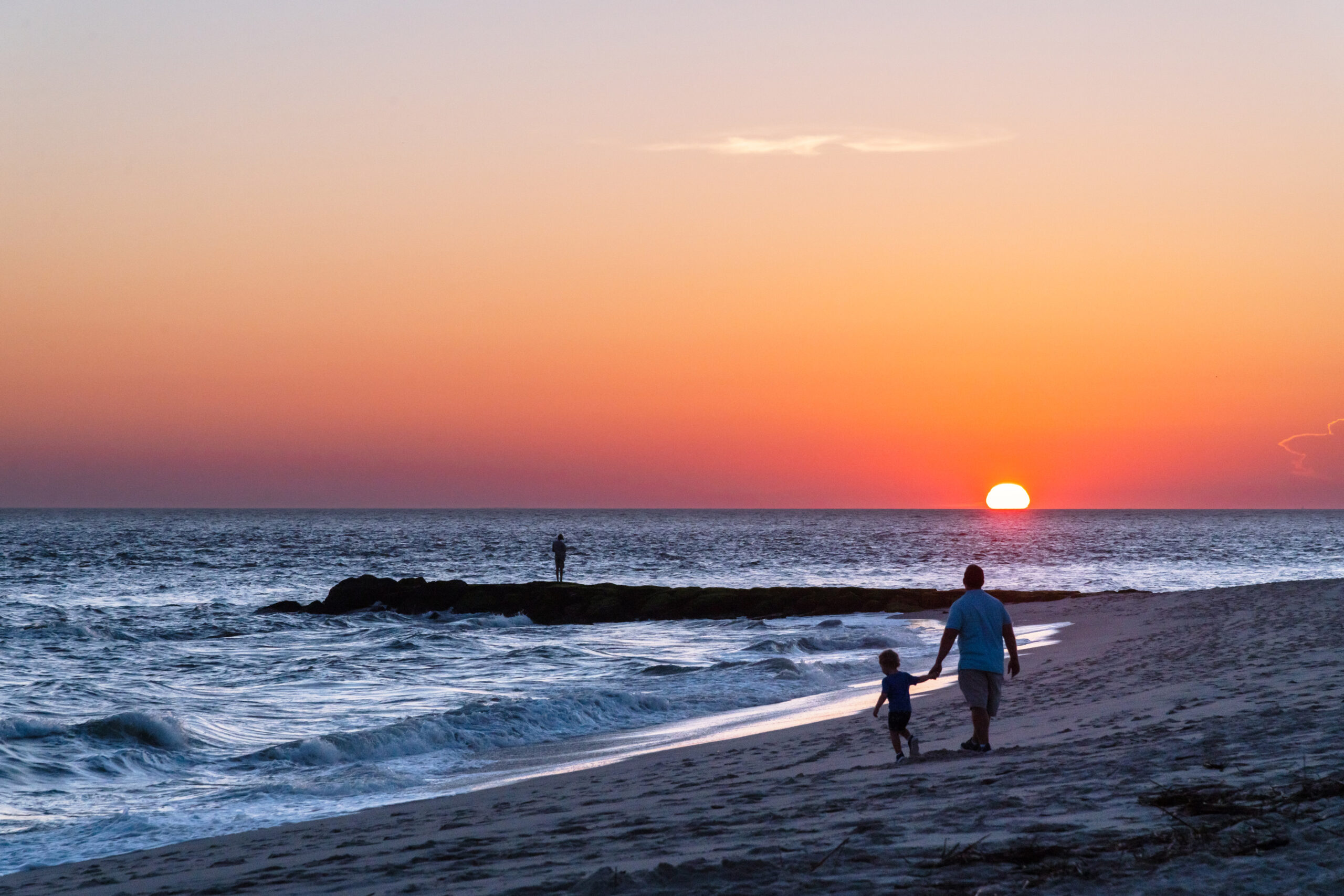 A father and son walking by the ocean on the beach as the sun sets below the horizon. The sky is clear and pink and orange. A person is out in the distance standing on the rocks leading out to the ocean.