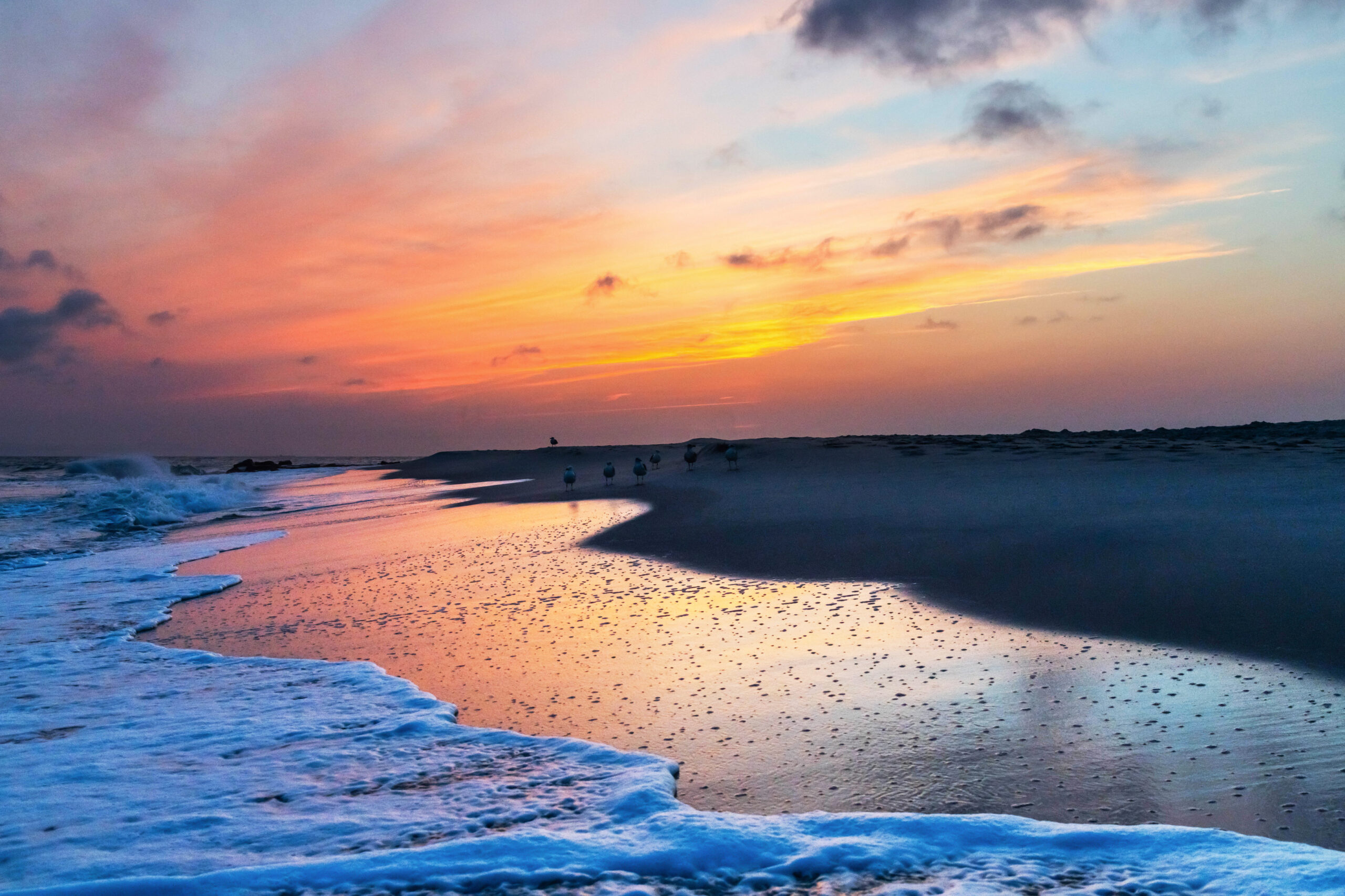 A colorful sky with pink, red, orange, yellow, and purple thin clouds. The ocean is rushing into the shoreline in the foreground, and the colors from the sky are reflected in the ocean.