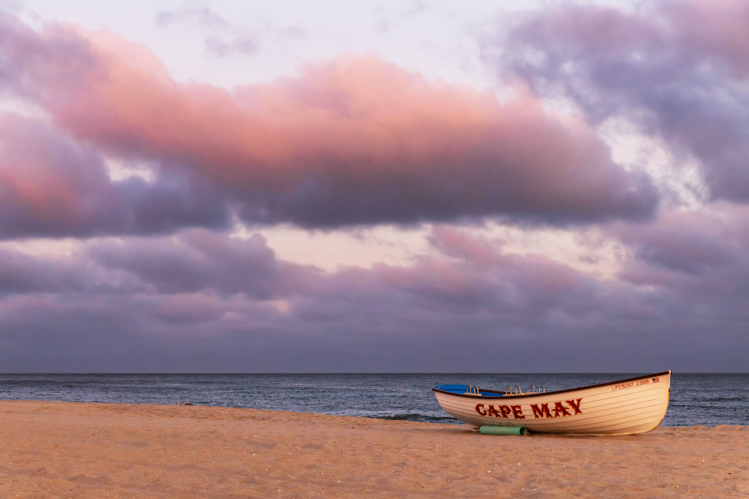 A Cape May lifeguard boat on the beach. The boat is white with Cape May written in red lettering. There are pink and purple puffy clouds in the sky.