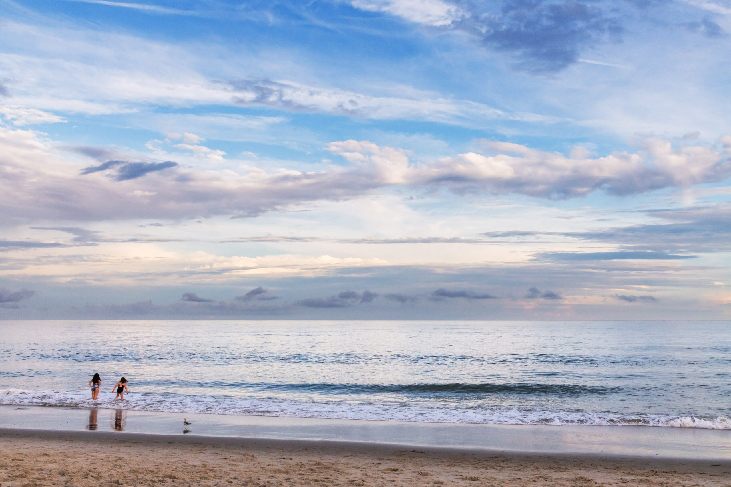 Thin wispy and fluffy clouds in a blue sky. The ocean is pale blue, and two people are going into the ocean.