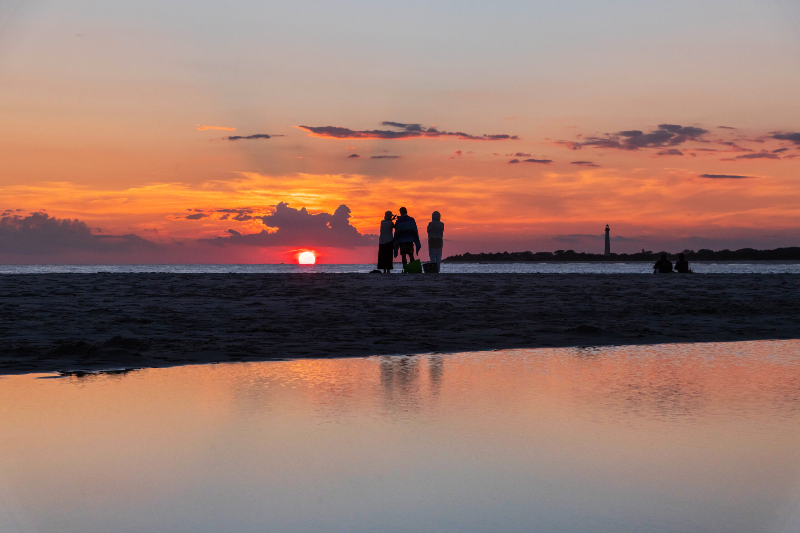 Three people standing at the edge of the beach watching the sunset with the Cape May lighthouse in the distance. The sun is bright pink, and there are purple and orange clouds in the sky. There is a body of water in the foreground, and the colors in the sky are reflected in the water.