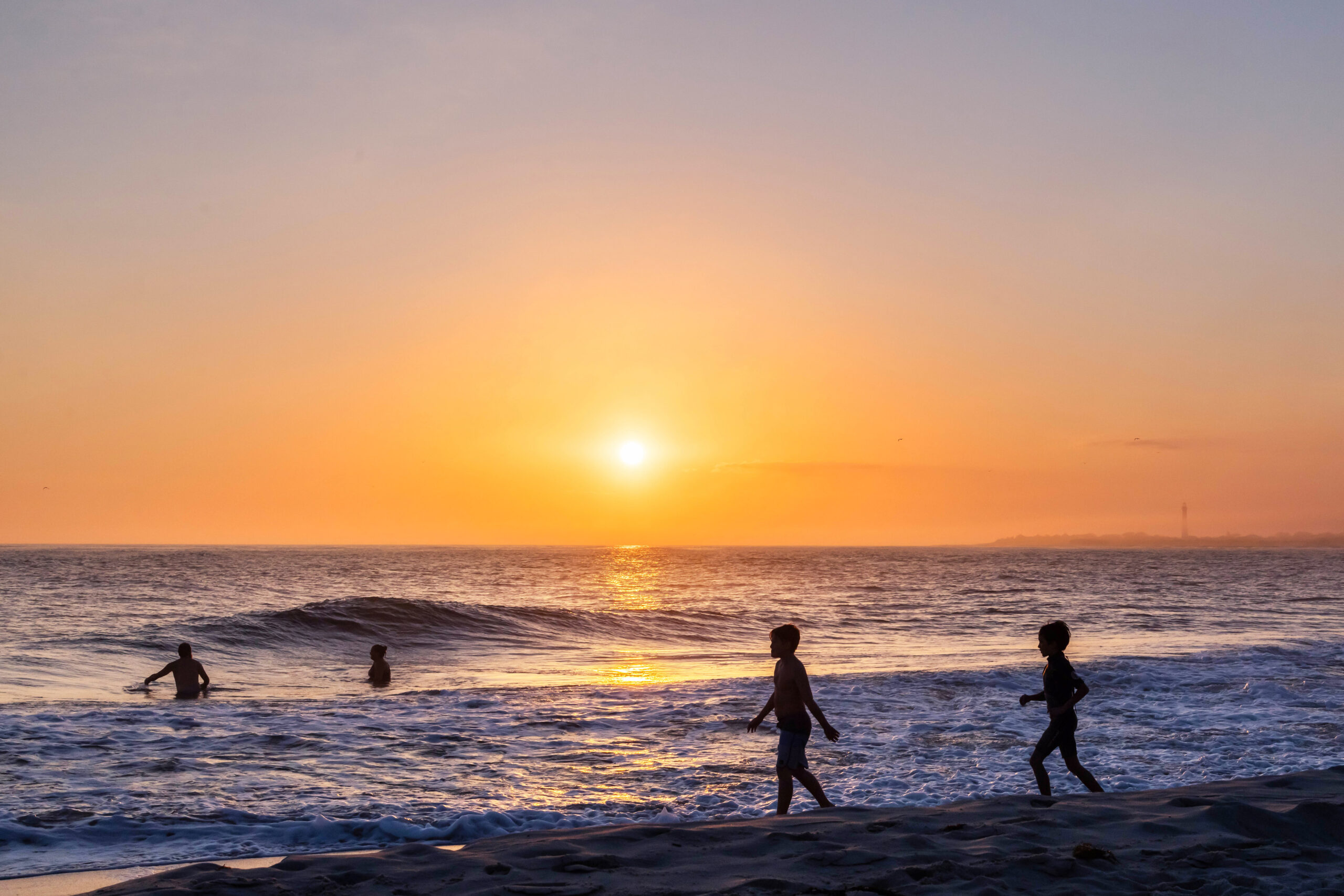 The sun setting at The Cove beach with a clear but hazy sky. Two people are swimming in the ocean, and two kids are running into the ocean. The Cape May Lighthouse is in the distance are partially obscured by fog.