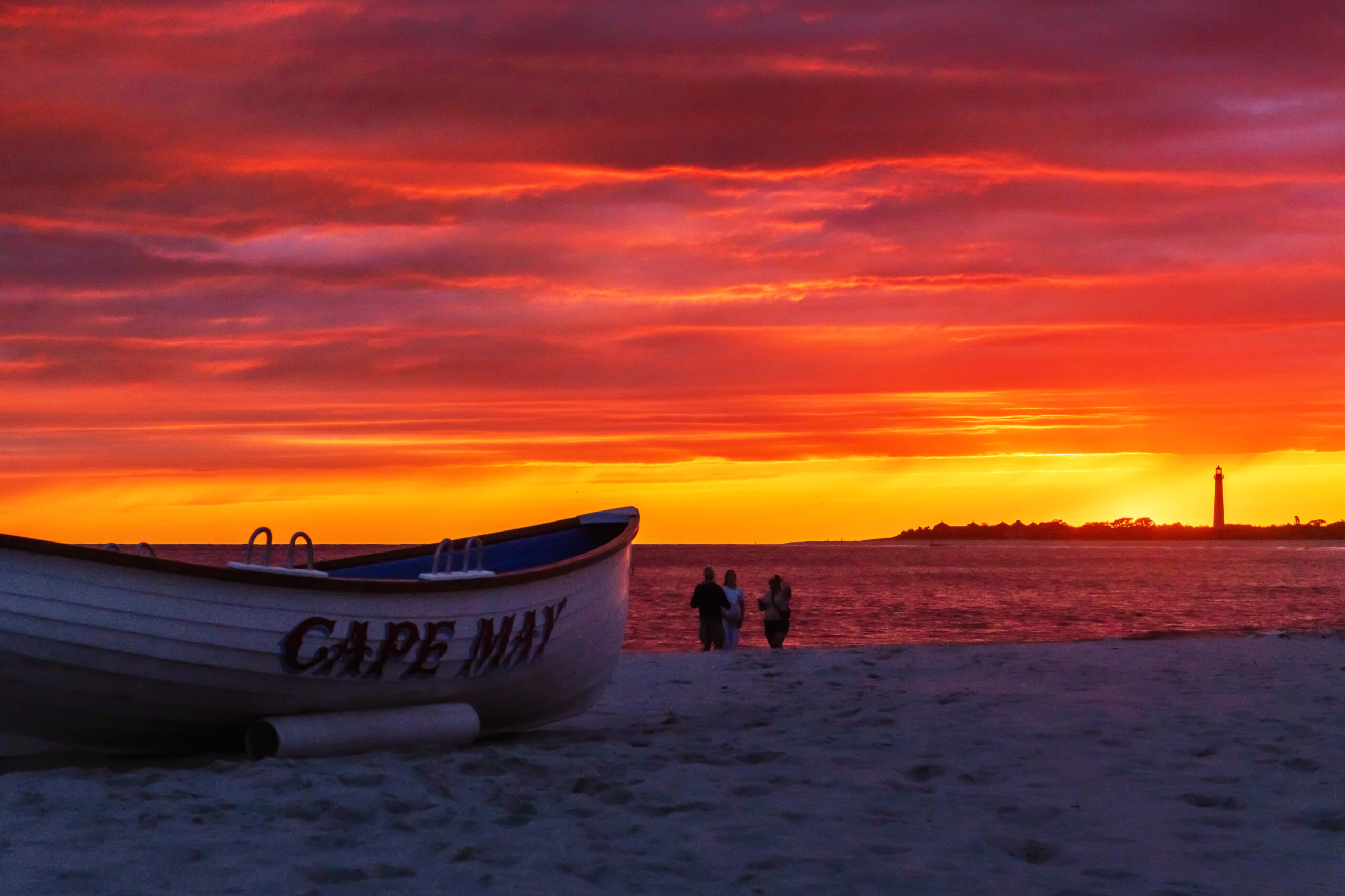 A Cape May lifeguard boat on the beach at sunset. There are brilliant red and orange clouds in the sky, and bright golden light at the horizon. The Cape May Lighthouse is in the distance.