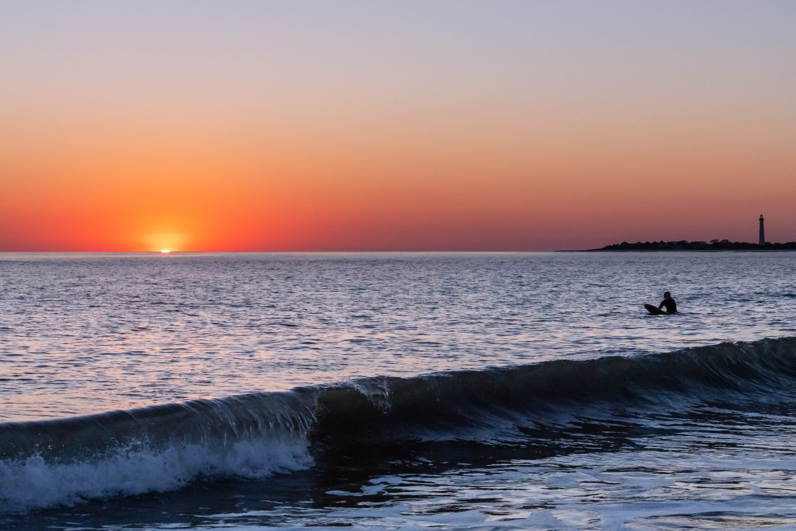 A wave crashing in the ocean as the sun sets behind the horizon in a clear sky. There is one surfer in the ocean, and the Cape May Lighthouse is in the distance.