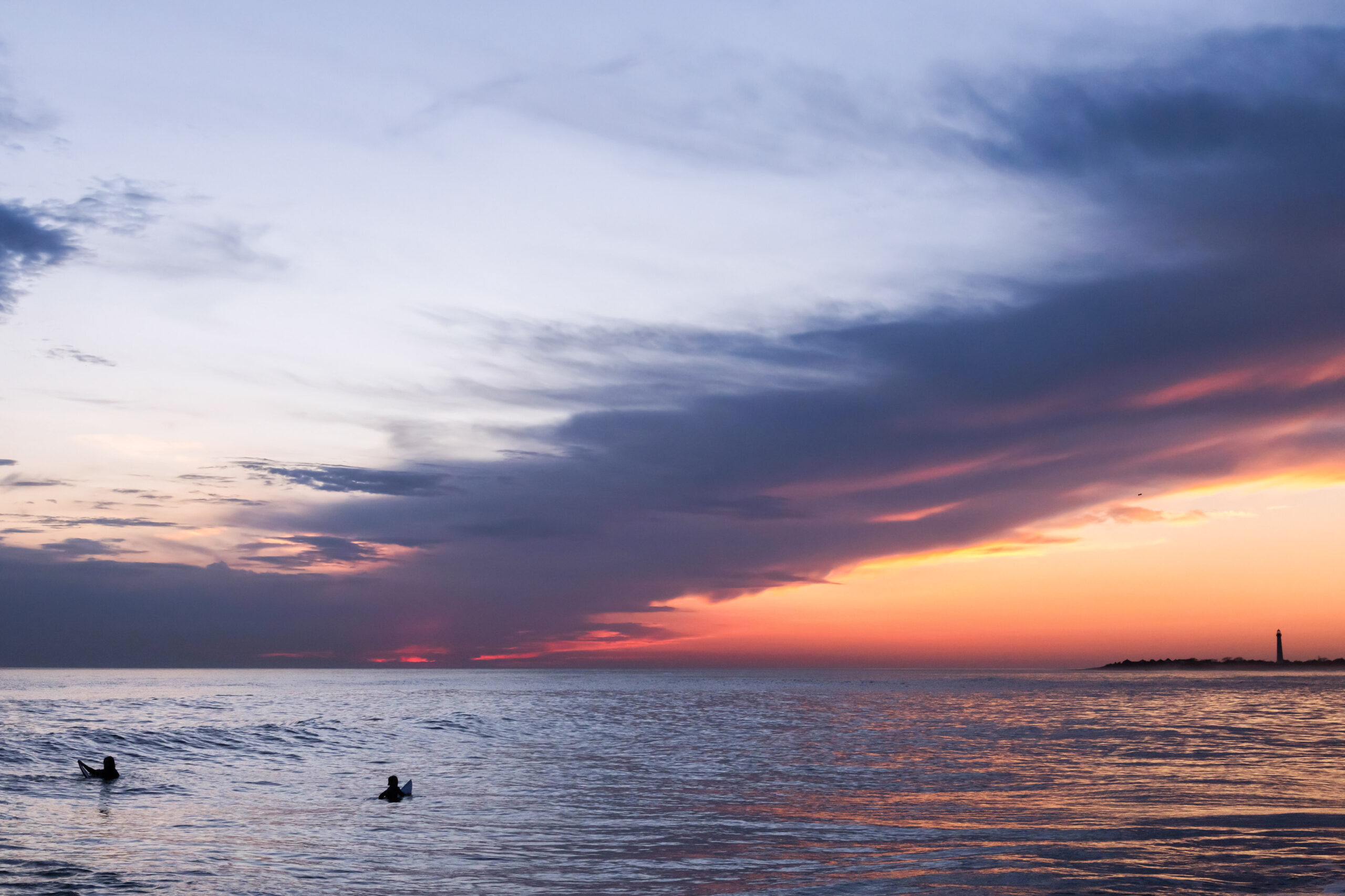 Two surfers in the ocean at sunset. There is a thin purple and pink cloud diagonally across the otherwise clear sky. Blues, purples, pinks, and oranges are reflected in the ocean. The Cape May Lighthouse is visible in the far right.