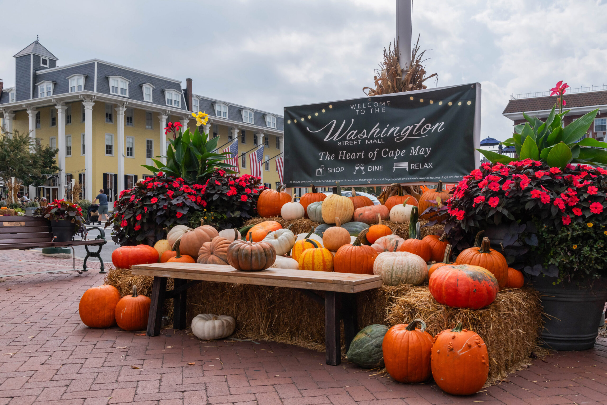 Piles of orange, yellow, green, and white pumpkins on haystacks with red flowers. The fall decor is in front of a sign that says "Welcome to the Washington Street Mall, The Heart of Cape May, Shop, Dine, Relax". Congress Hall is in the background.