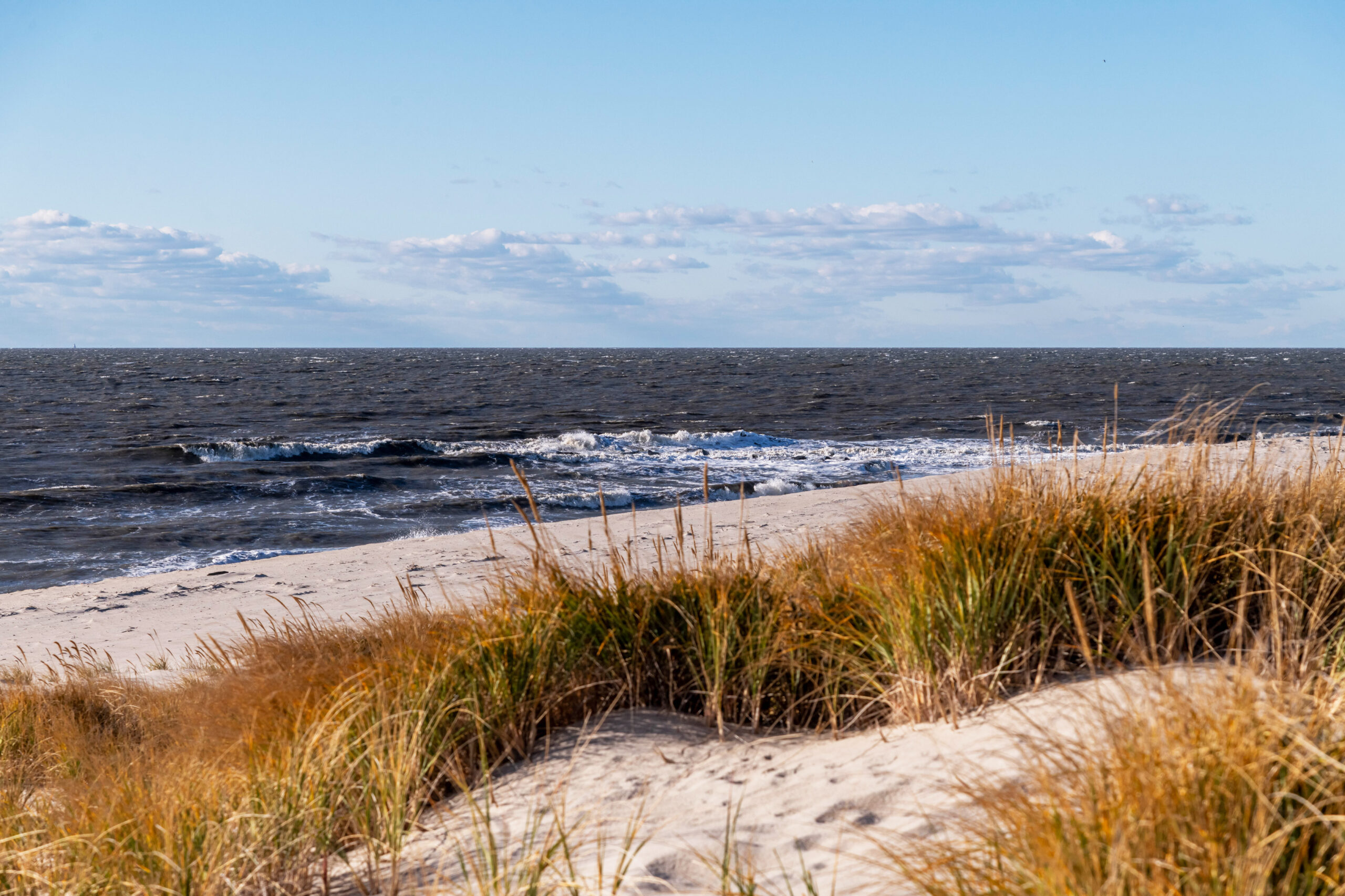 Orange dunes in sand in the foreground of the photo, with waves crashing in the ocean in the background. The ocean looks choppy with white caps. There are a few puffy white and gray clouds in the blue sky in the distance.