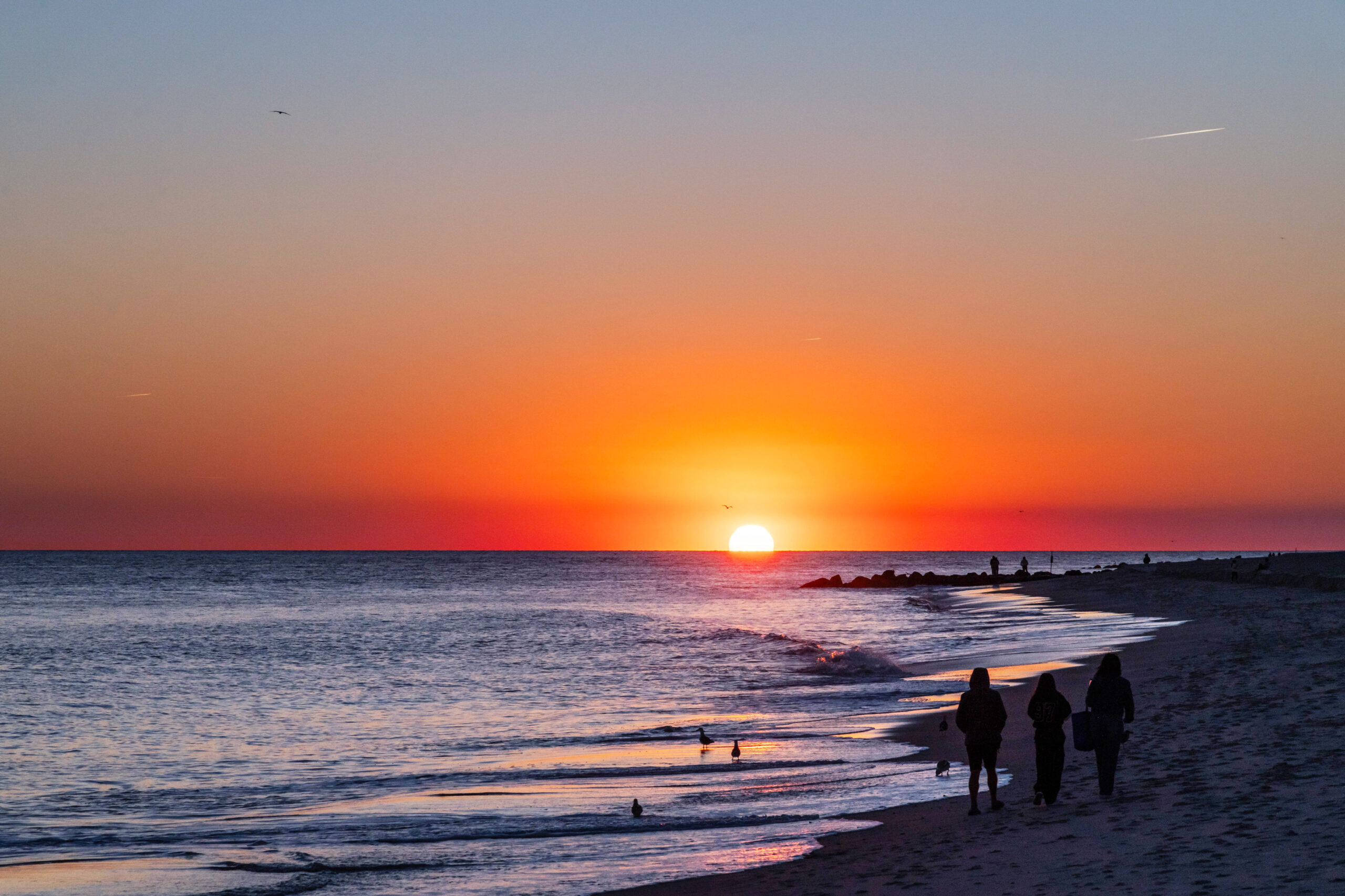 Three people walking on the beach at sunset. There is a clear blue and orange sky, and some birds are in the ocean.