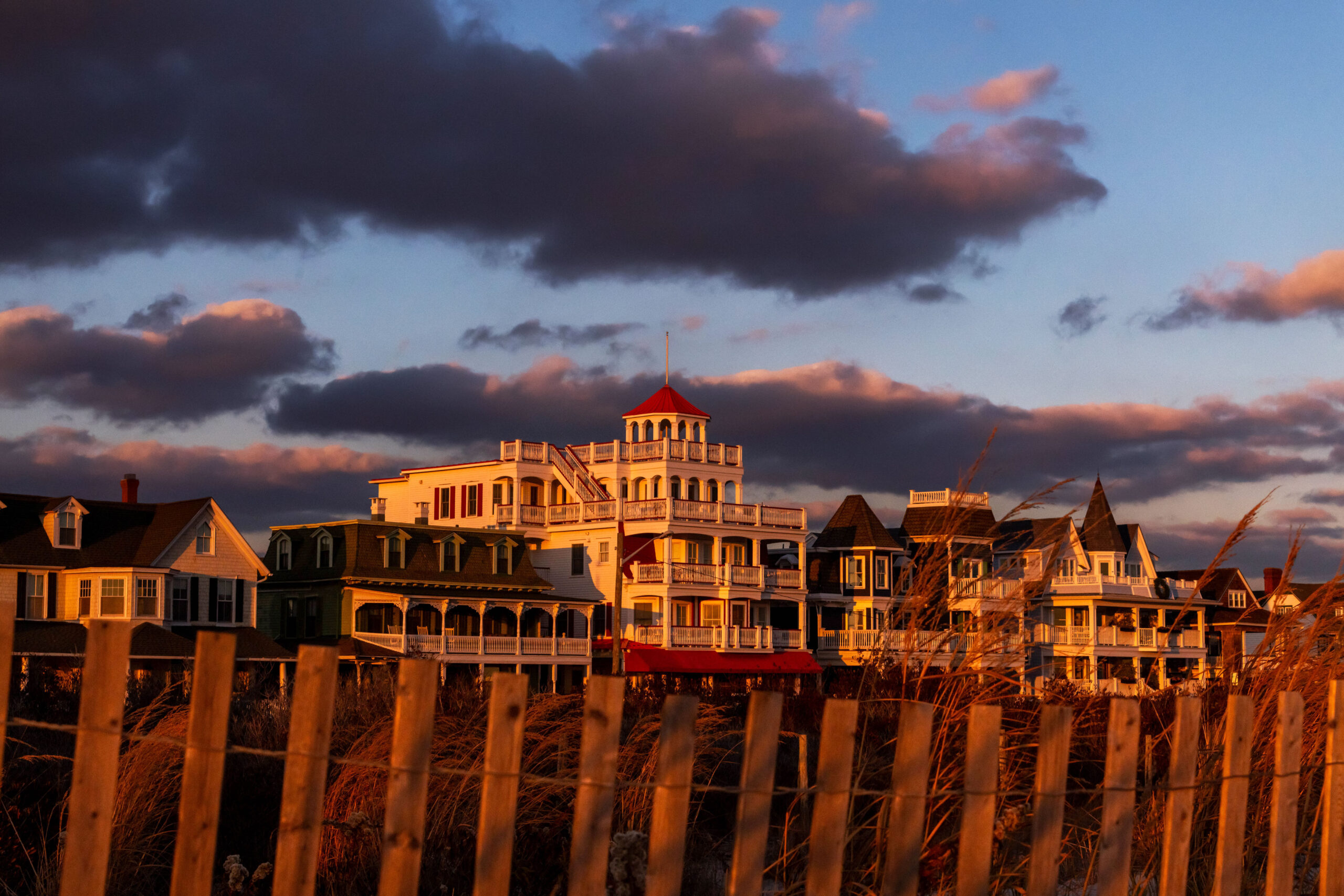 Purple clouds behind Victorian houses at sunset. There are orange dunes and a beach fence in front of the houses. Sunset light is shining on the houses and the dunes.