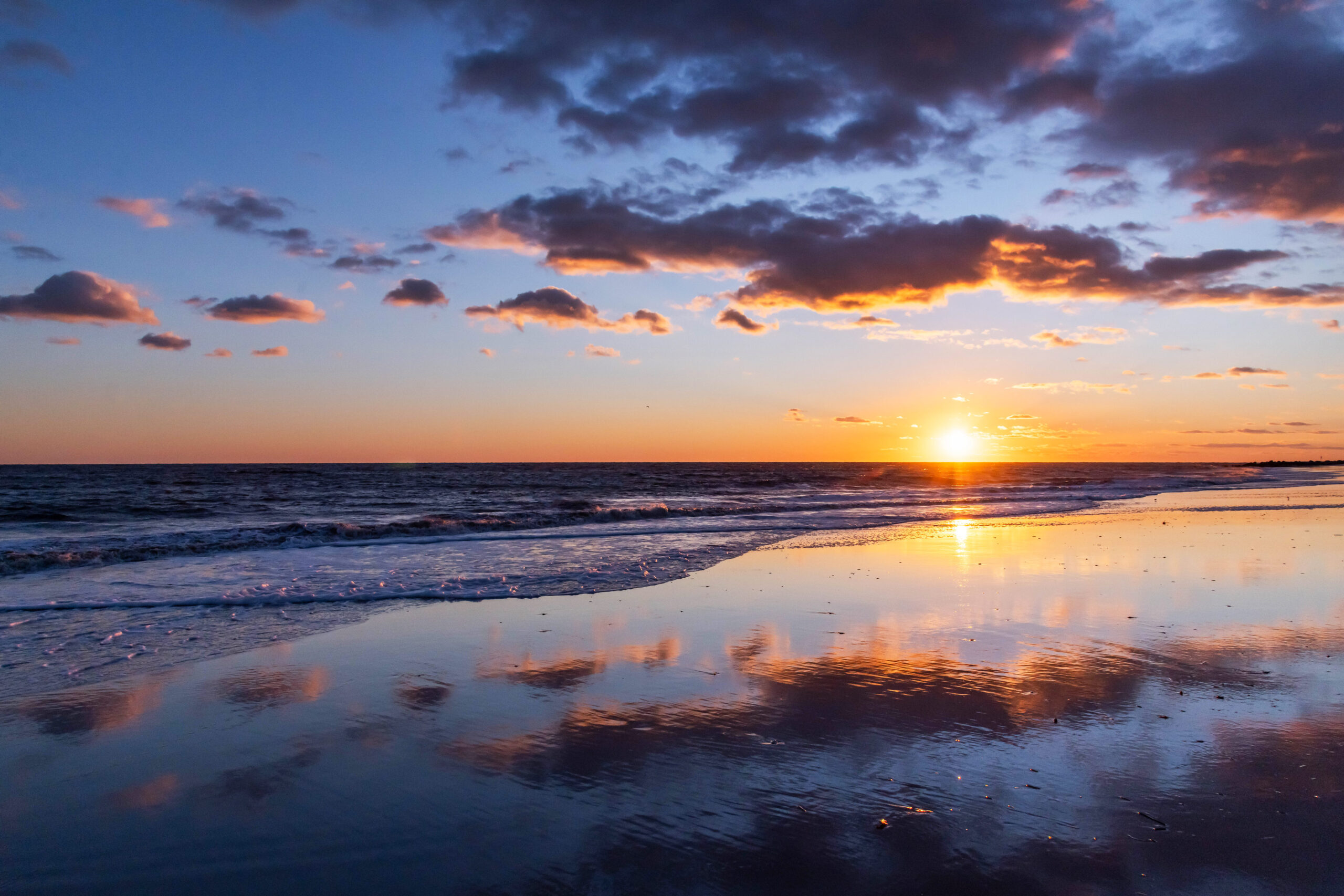 A wide view of the sunset. Puffy purple and orange clouds are in the sky and are reflected the blue ocean and sand.