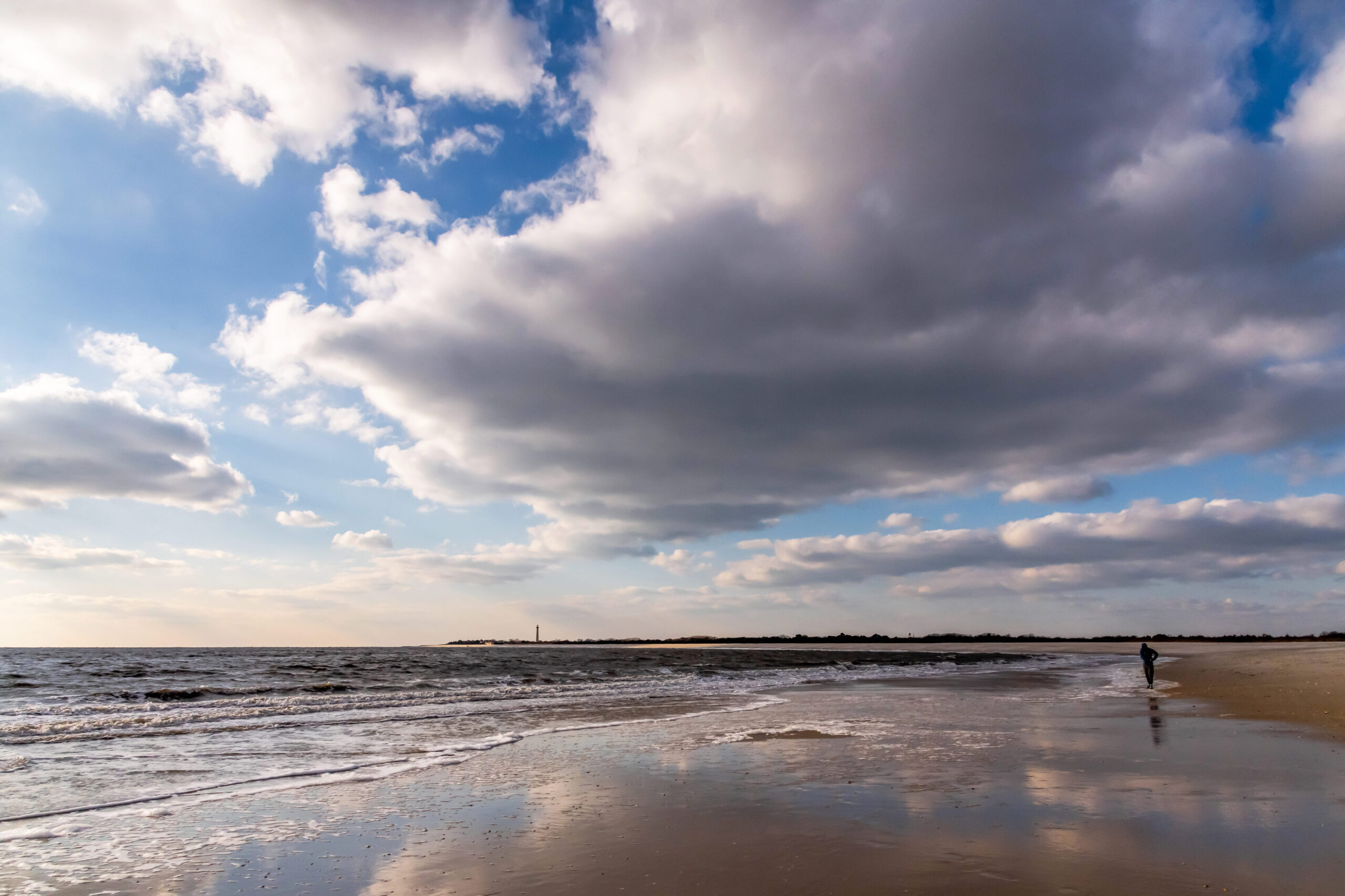 Wide angle view of the ocean, beach, and sky at The Cove beach. There are big white puffy clouds in a blue sky, and the clouds are reflected in the sand and ocean. Someone is walking on the beach, and the Cape May Lighthouse is in the distance.