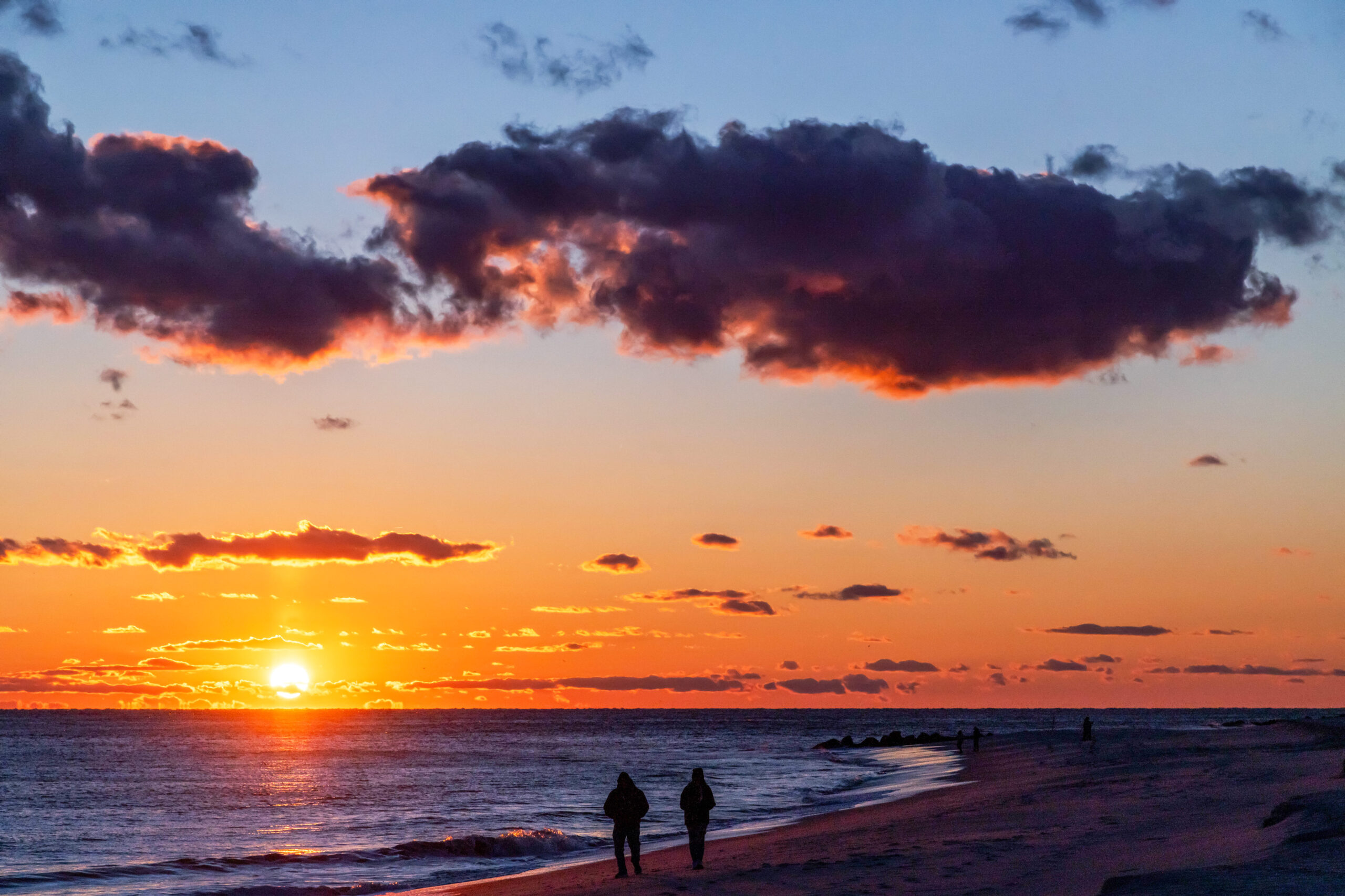 A wide view of the sun setting at the beach. There are a few puffy purple and orange clouds in an otherwise clear sky. Two people are walking on the beach by the ocean.