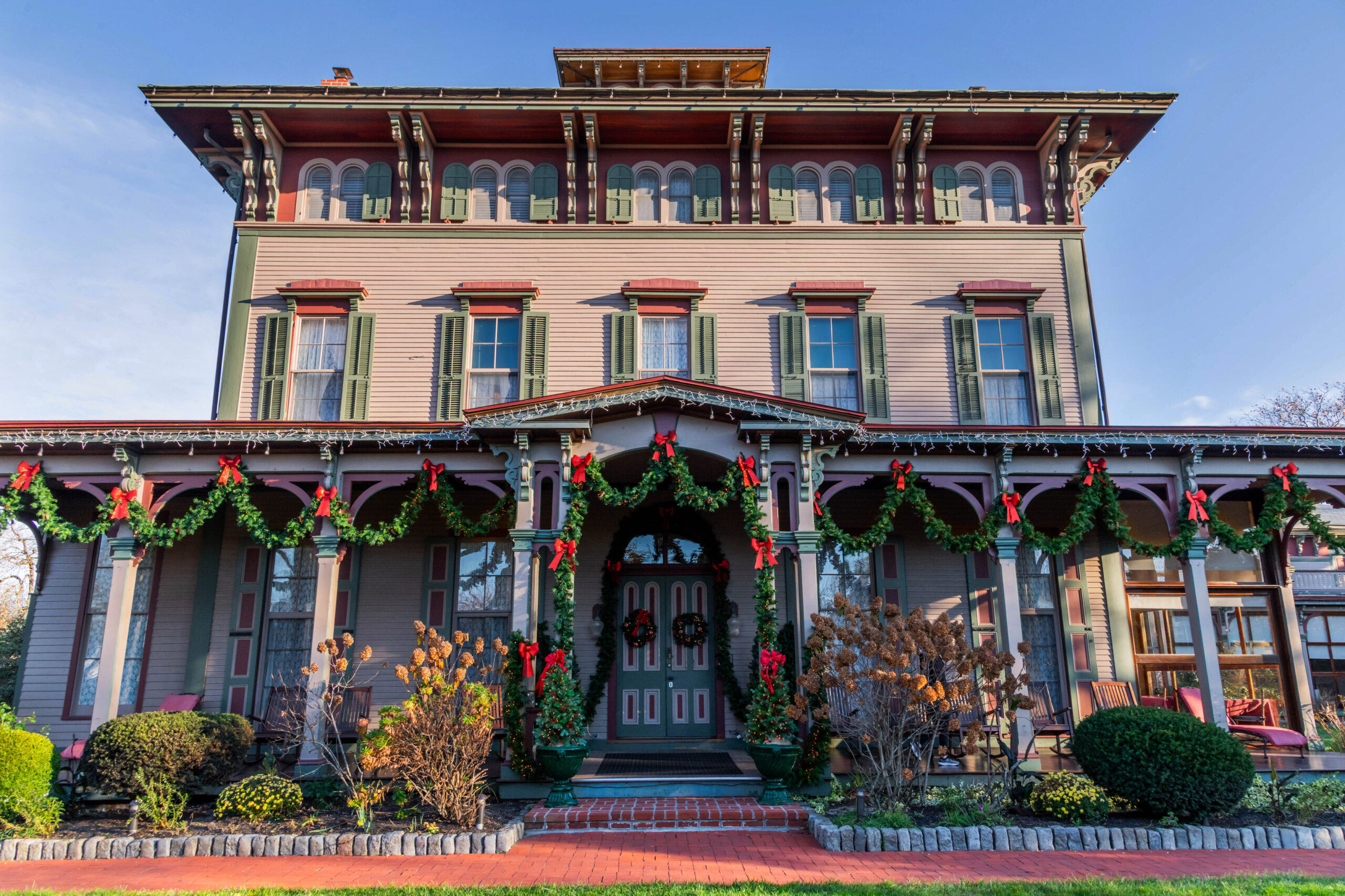 A wide view of a Victorian styled building decorated with green garland and red bows. The garland is hanging from the top of the front porch and in front of the door. The building is light pink with olive green shudders and a darker pink trim. The sun is shining on the building, and there is a clear blue sky.