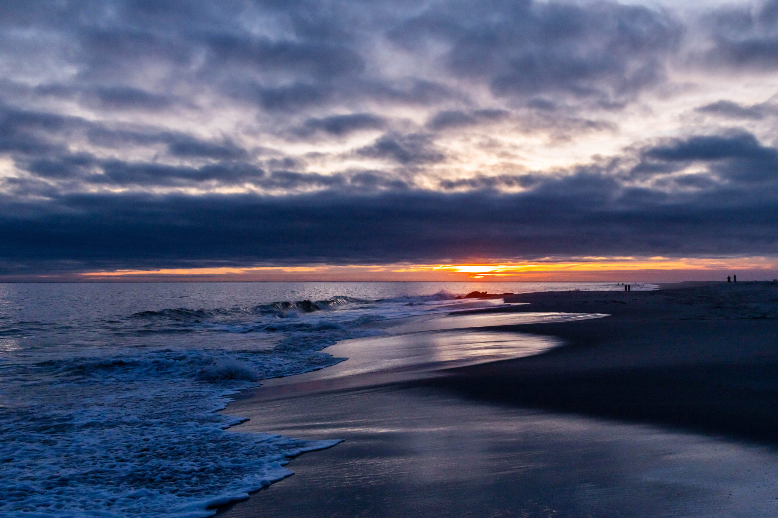 A wide view of the sun setting behind clouds with small waves crashing in the ocean. There is a stripe of orange at the horizon, and there is a dark purple blue cloud in the sky breaking up. The clouds are reflected in the shoreline and sand.