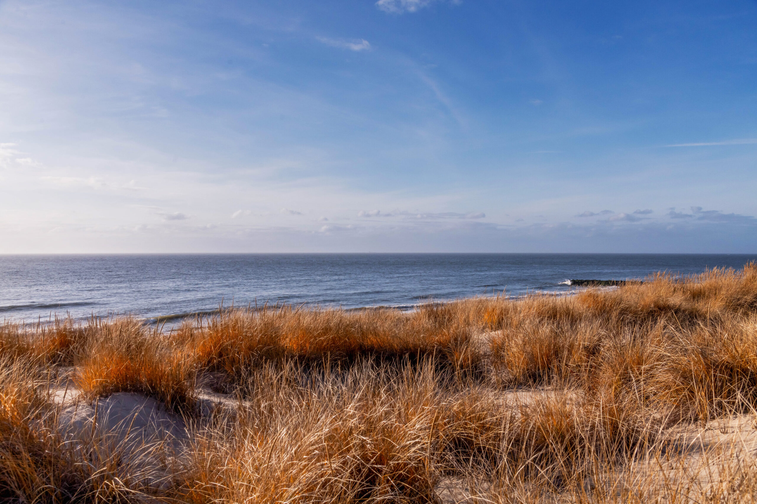 A wide view of beach dunes, the ocean, and a blue sky with a few thin white clouds. The dunes are orange and there are a few waves crashing in the ocean.