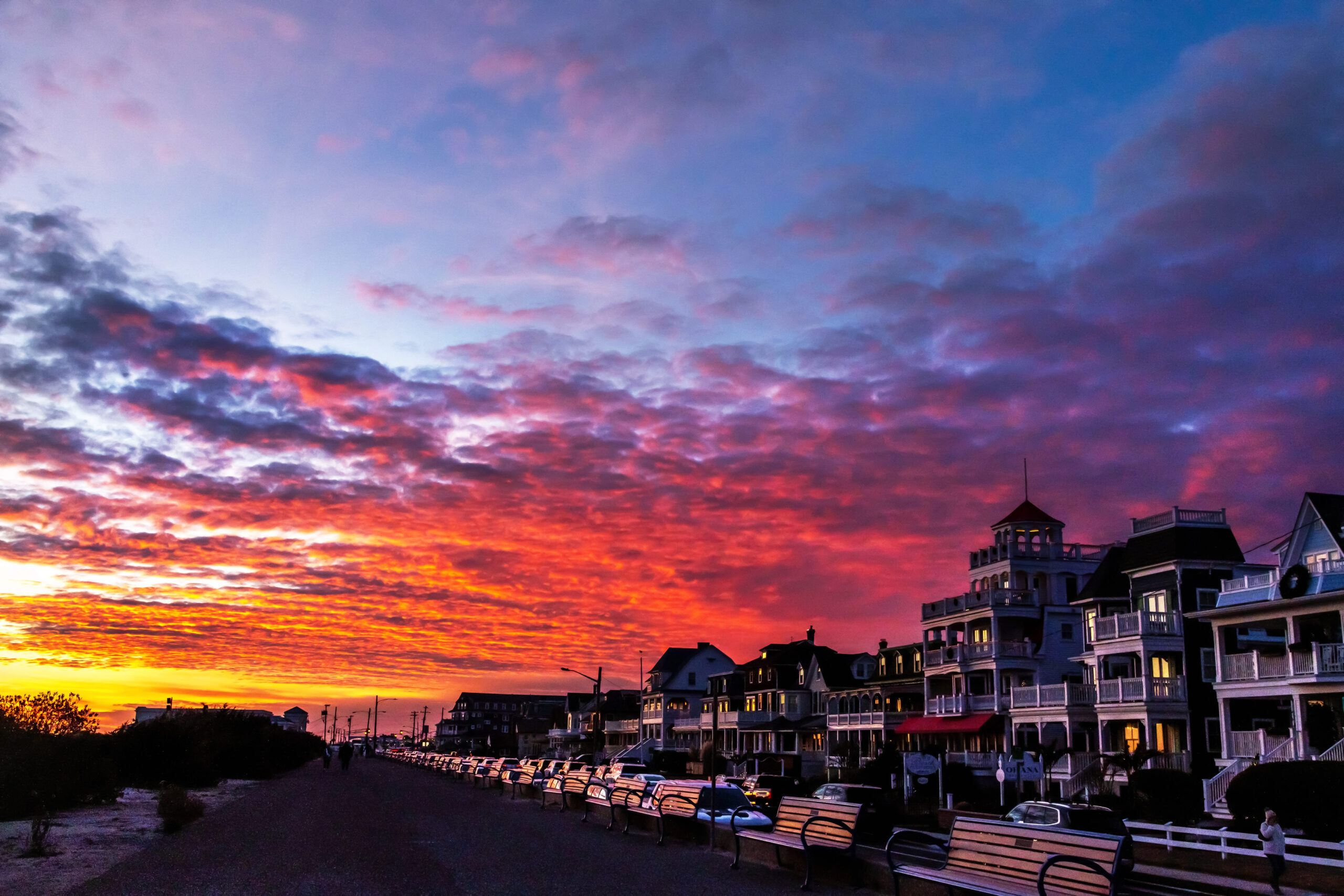 Orange, pink, and purple clouds reaching across the sky at sunset. The Victorian houses along Beach Ave are pictured behind the benches on the promenade.