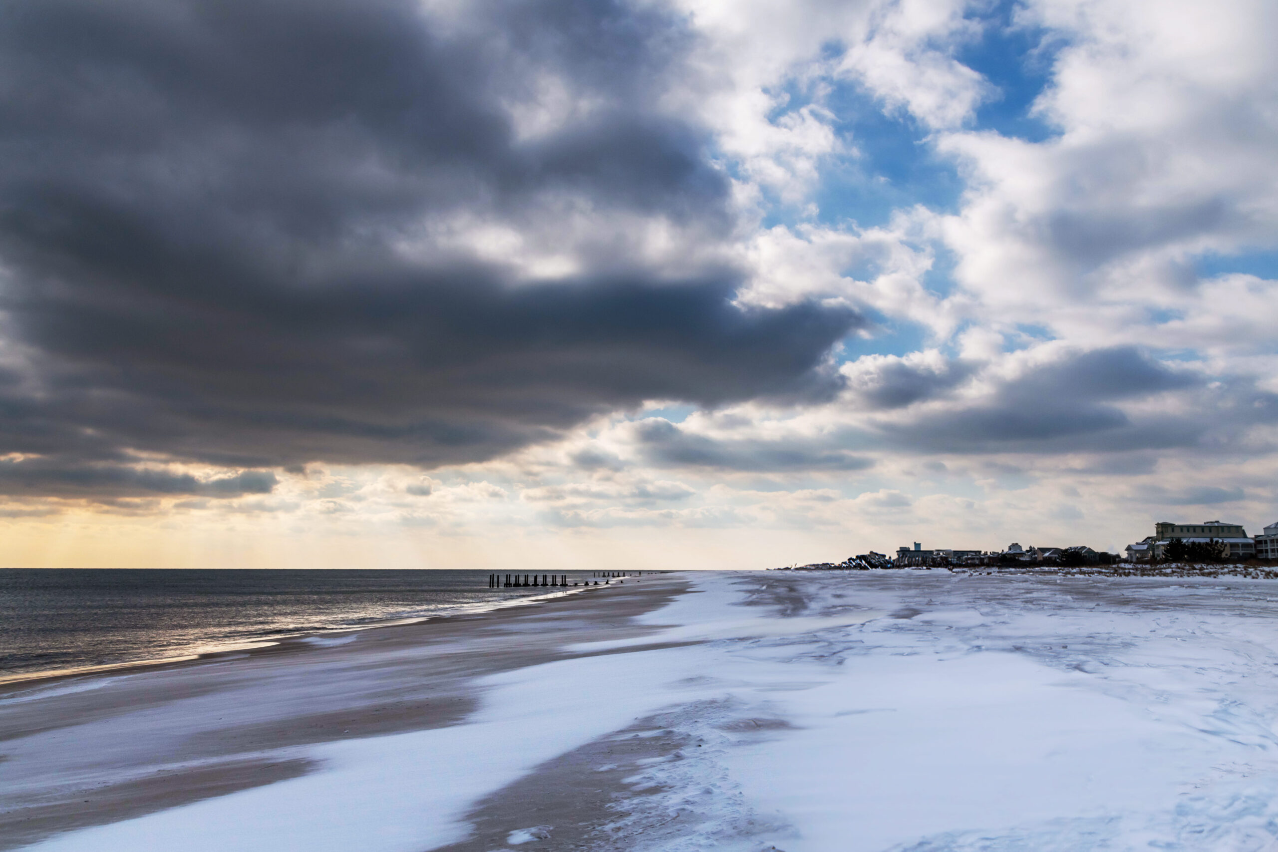 A wide view of snow on a beach with the sun shining behind big puffy dark clouds. There is blue sky behind the clouds, and yellow light is shining on the ocean.