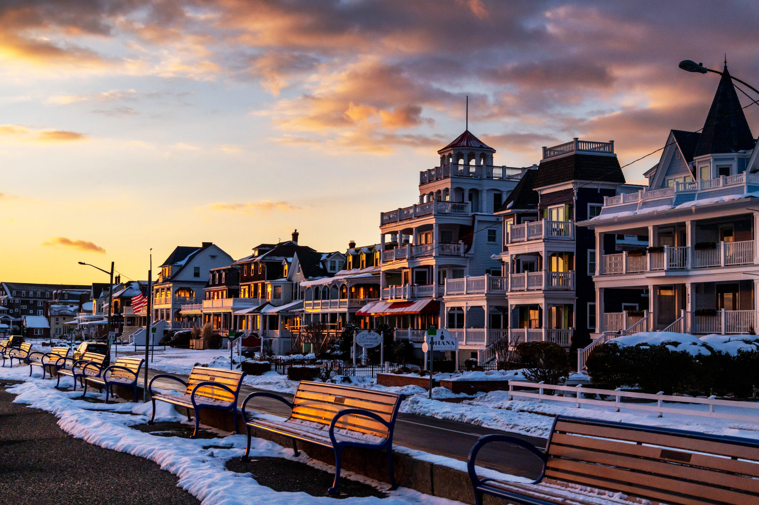 A wide view of the benches on the promenade and Victorian houses on Beach Ave at sunset. There is melting snow on the benches and on the houses. There are a few orange, pink, and purple clouds in an otherwise clear yellow and blue sky.