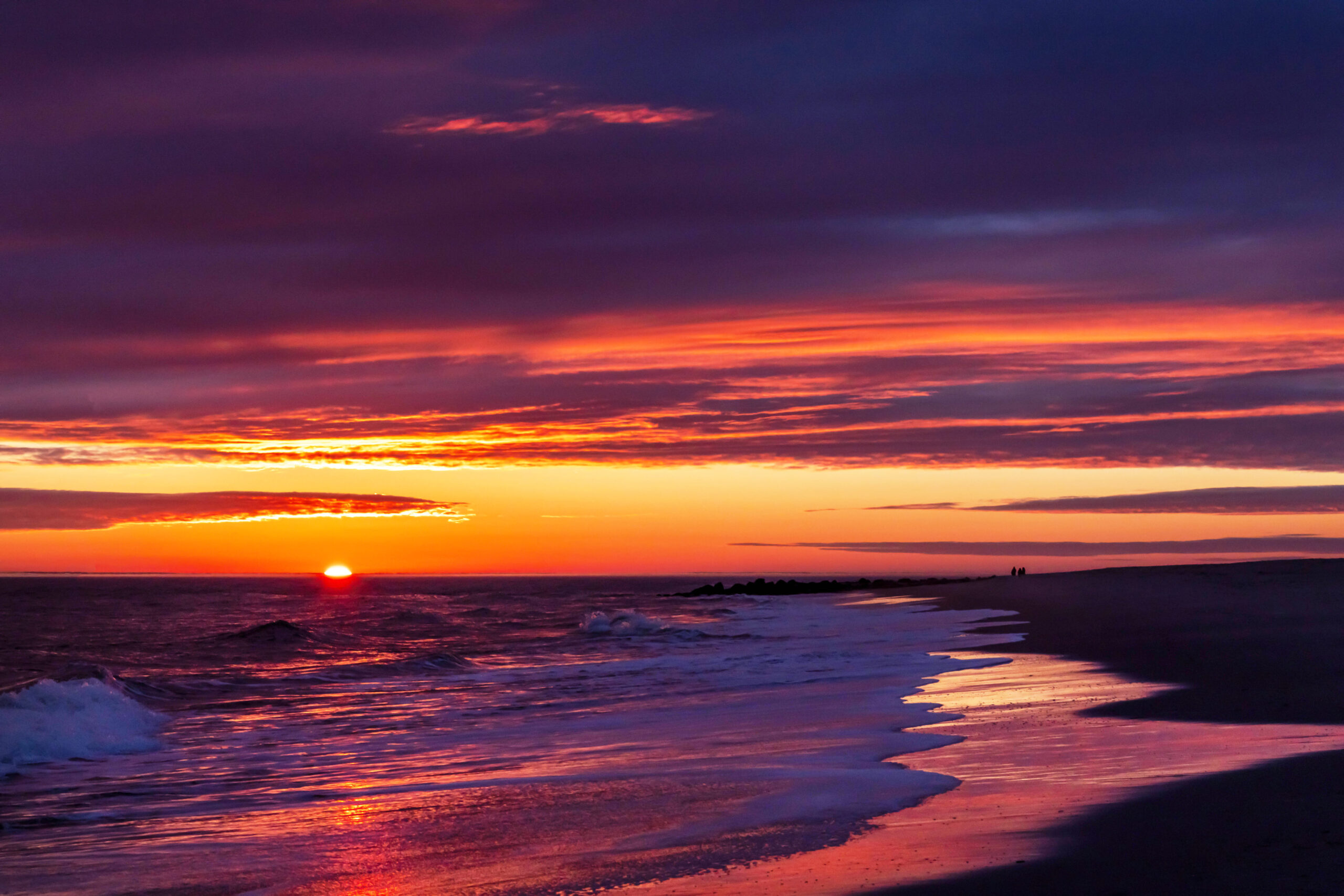 A wide view of a colorful sunset at the beach. There are dark purple clouds in the sky with orange and pink streaks of color. The sky is clear at the horizon and bright orange. The sun is half set behind the horizon. Waves are coming into the shoreline, and the sky is reflected in the sand and ocean.