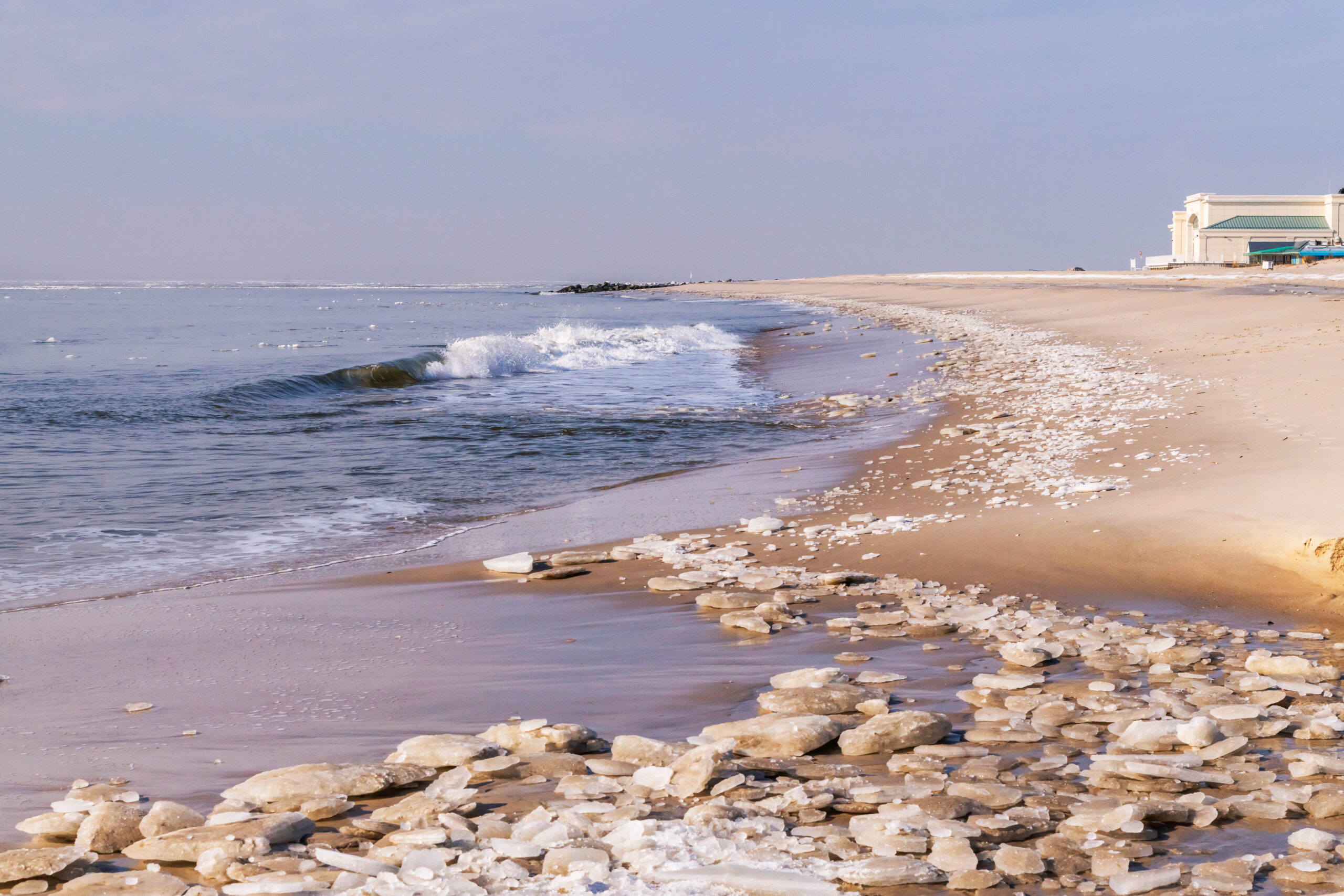 A wide view of the beach and ocean with blocks of ice along the shoreline and some ice in the distance in the ocean. The Cape May Convention Hall is in the distance, and a wave is crashing in the ocean.