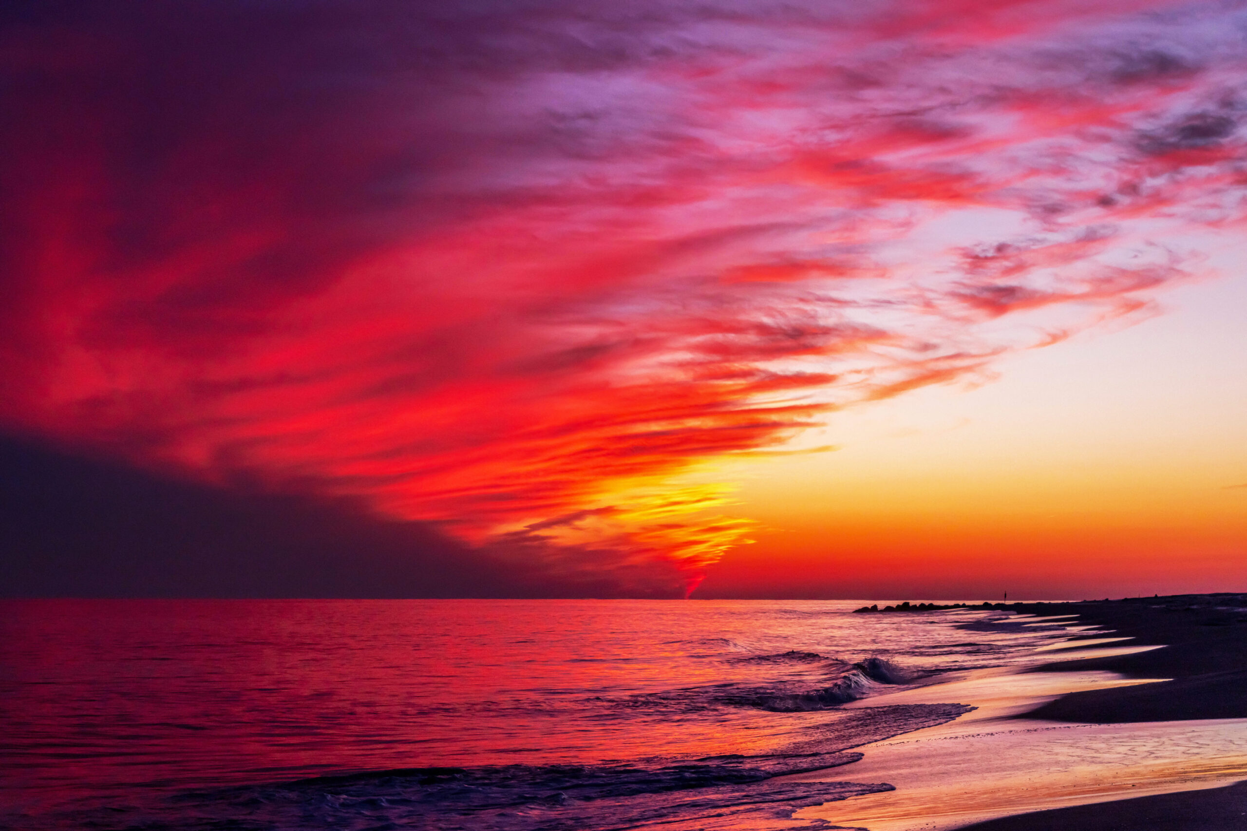 A wide view of colorful sunset at the beach. There is a brilliant red, purple, orange, and yellow cloud in taking up half of the sky. There is clear sky behind it and to the right. The brilliant colors are reflected in the ocean and the shoreline.