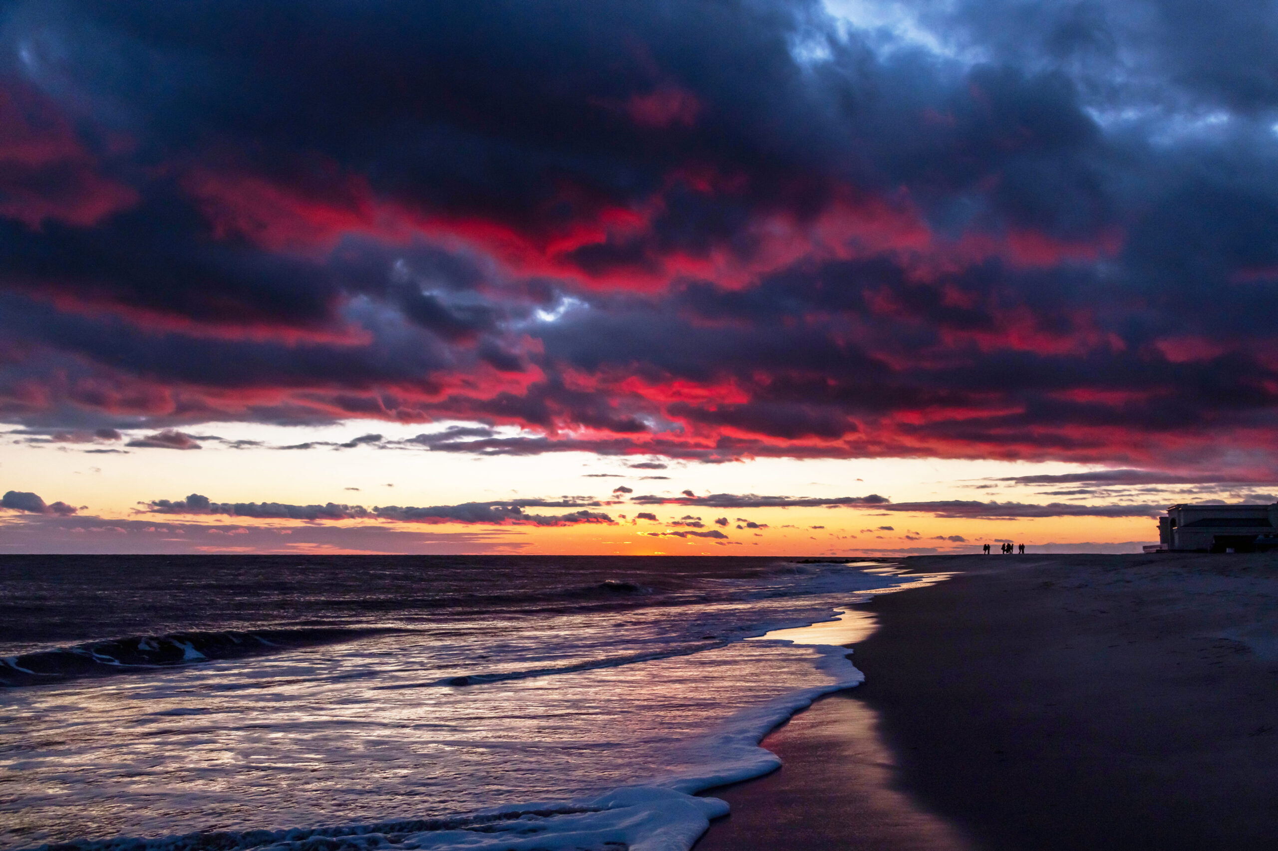 A wide view of the beach, ocean, and sky just after sunset. There are dark purple and pink clouds in the sky. At the horizon the sky is clear and orange. The ocean looks dark with some bright light reflecting from the sky. There is a wave coming into the shoreline, and Convention Hall and a few people are in the distance on the beach.