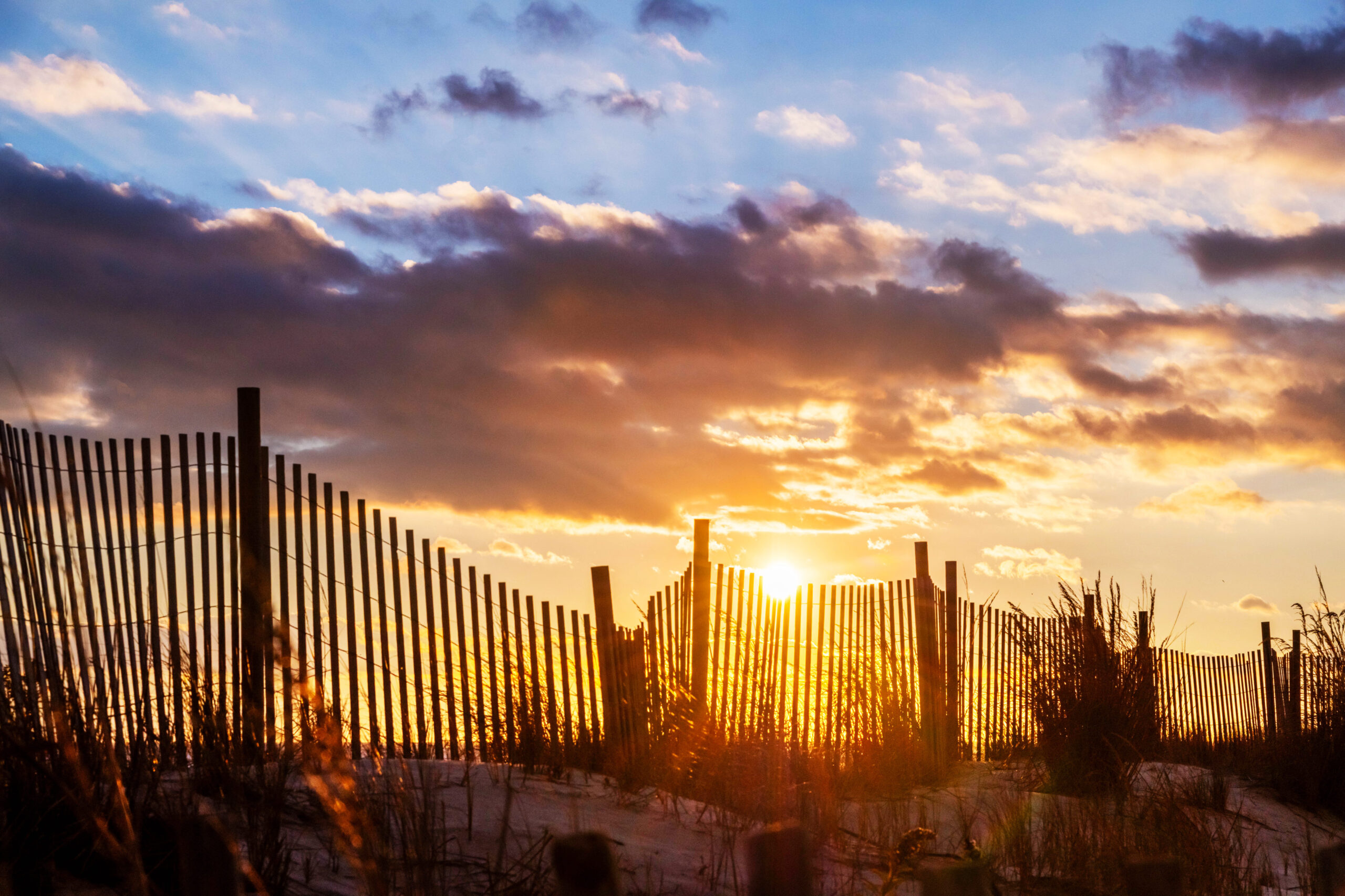 A wide view of the sun setting behind a beach dune fence, with some dunes in the foreground blurred out and puffy clouds in the sky. The clouds are purple, and the sky is blue and gold. The dunes and fence look dark and are silhouetted.