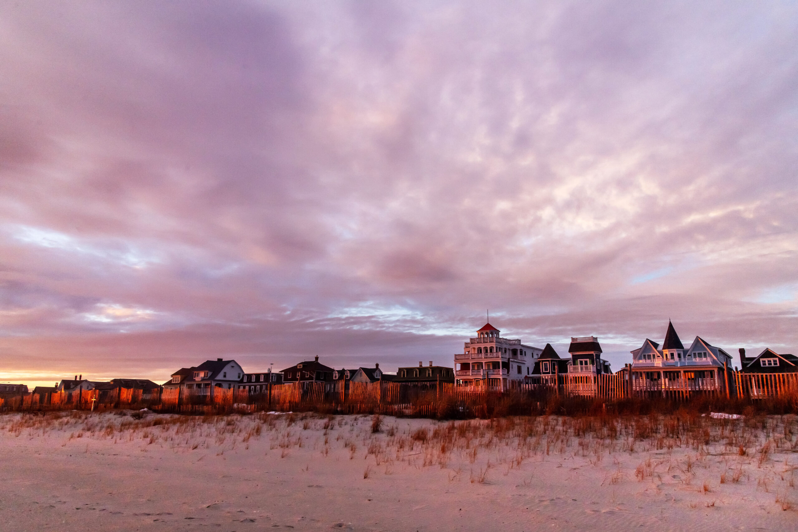 A wide view of the Victorian houses along Beach Ave from the beach. There are thin pink and purple colorful clouds in the sky. Orange light from the sunset is shining on the houses and the beach.