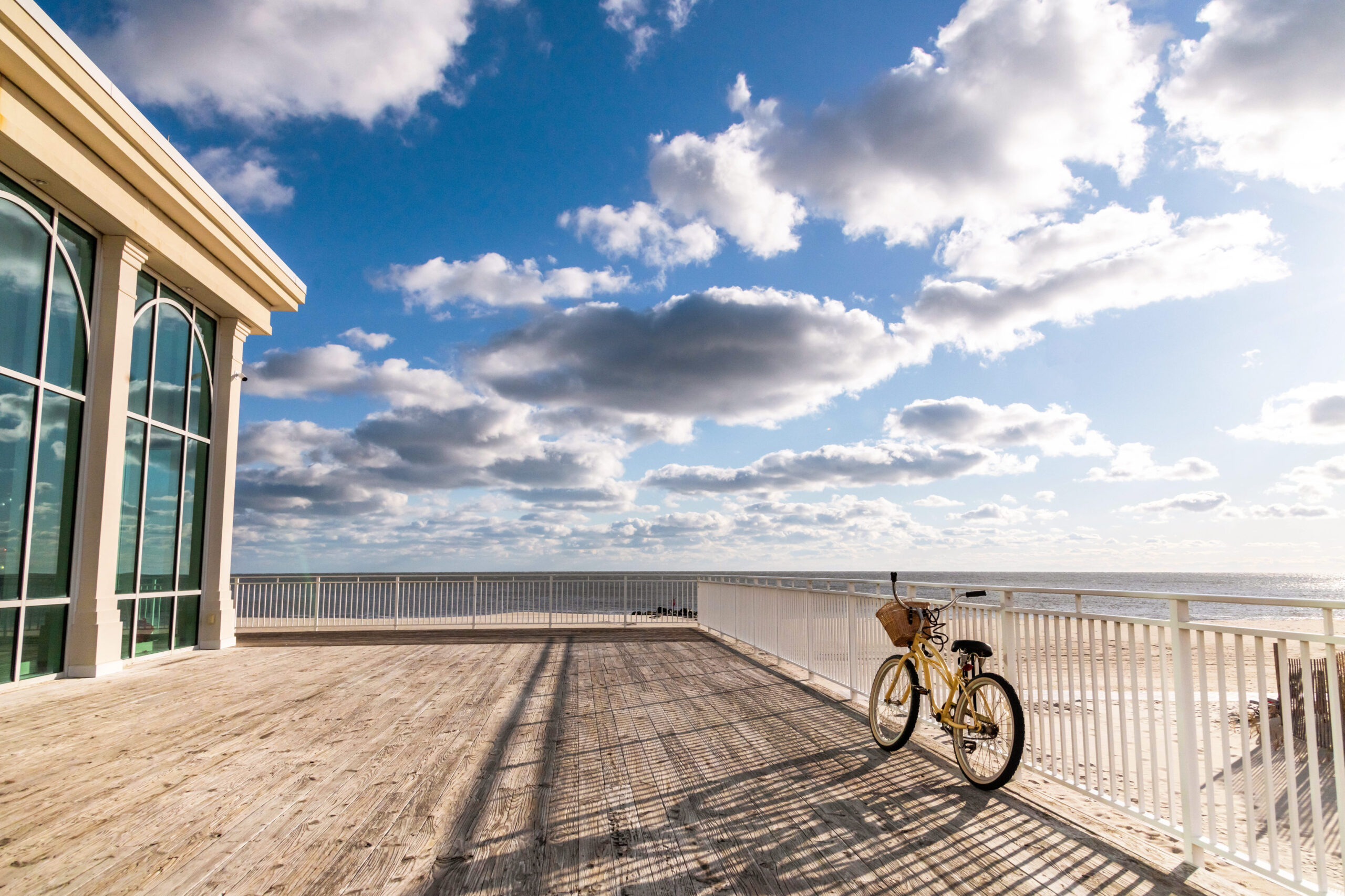 A wide view of the deck at Convention Hall. A yellow bike is resting against the railing on the right. The sun is shining bright and casting shadows on the deck. The side of Convention Hall with floor to ceiling windows is pictured on the left. There is a clear blue sky with puffy white clouds, and the beach and ocean can be seen in the background behind the deck.