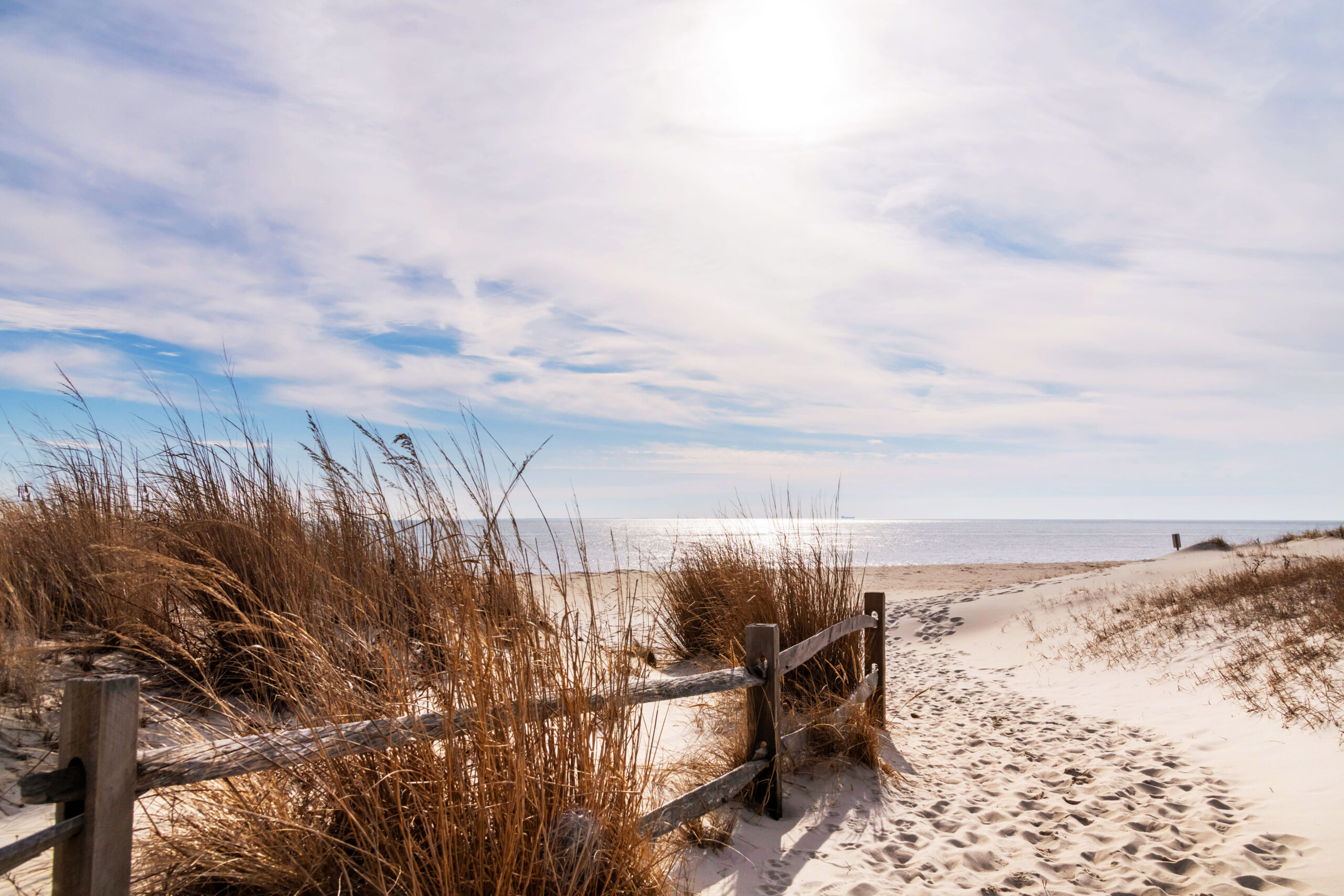 A wide view of a path leading to the beach. There is a fence with beach dunes. There are thin white clouds in a blue sky.