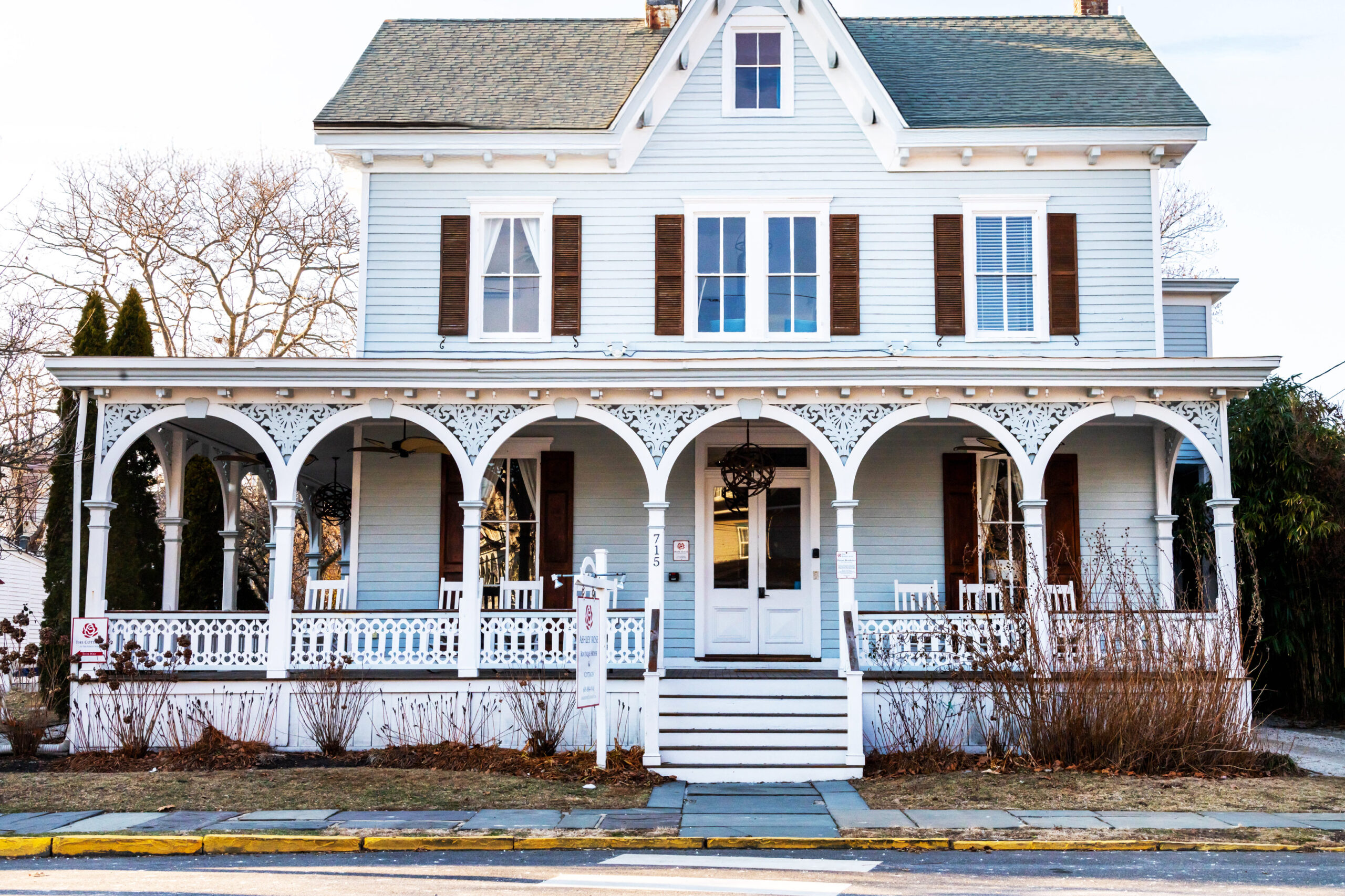 A wide view of a Victorian house. The house is pale light blue with dark brown shudders. There are white rocking chairs on the front porch. The sun is lightly shining on the house.