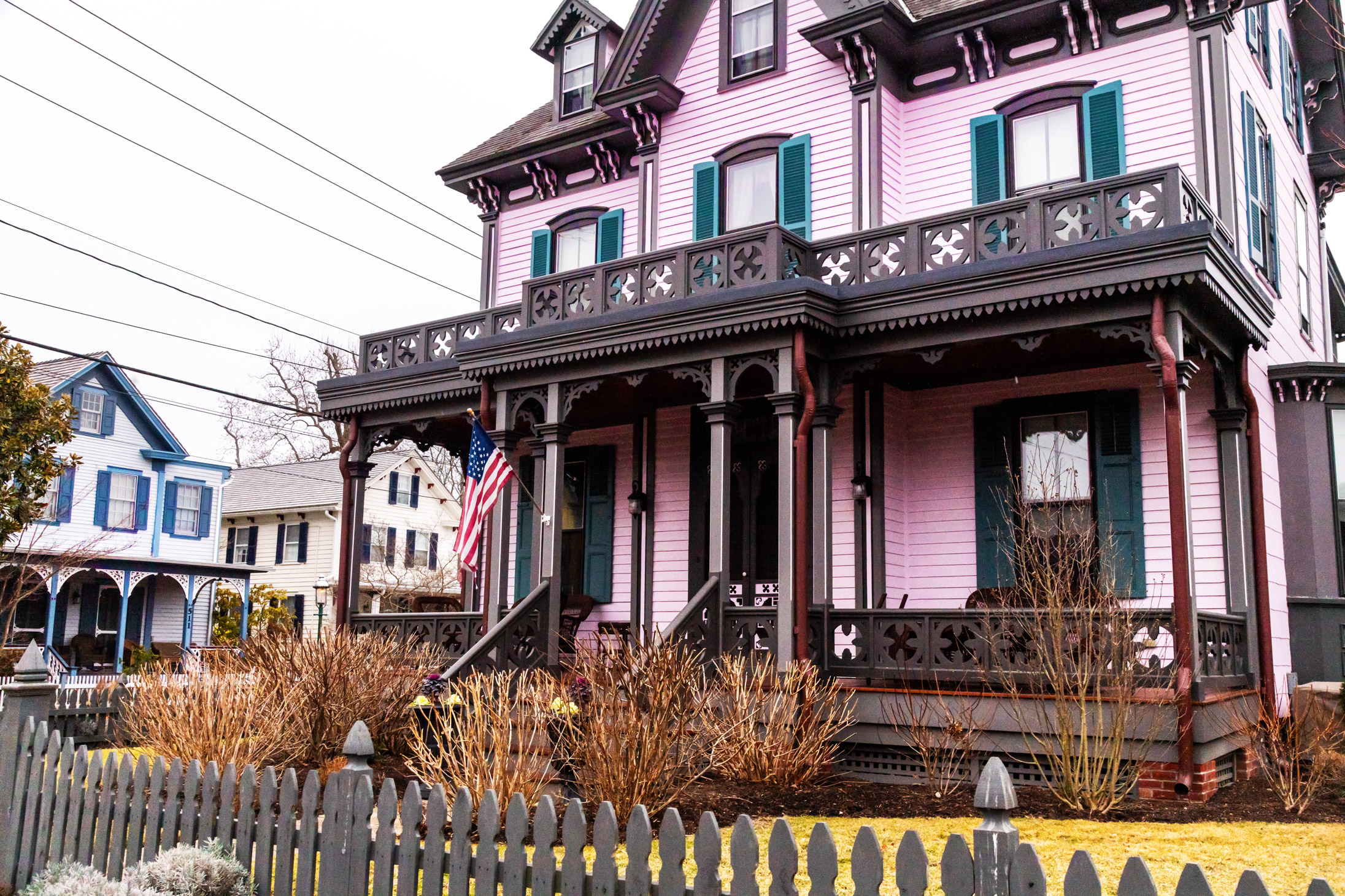 A wide angled view of a periwinkle Victorian house with teal shutters and brown railings and accents. There is an American Flag hanging off of the front porch. Gray clouds are in the sky, and a blue Victorian house is in the background. A brown fence is in the foreground of the image.