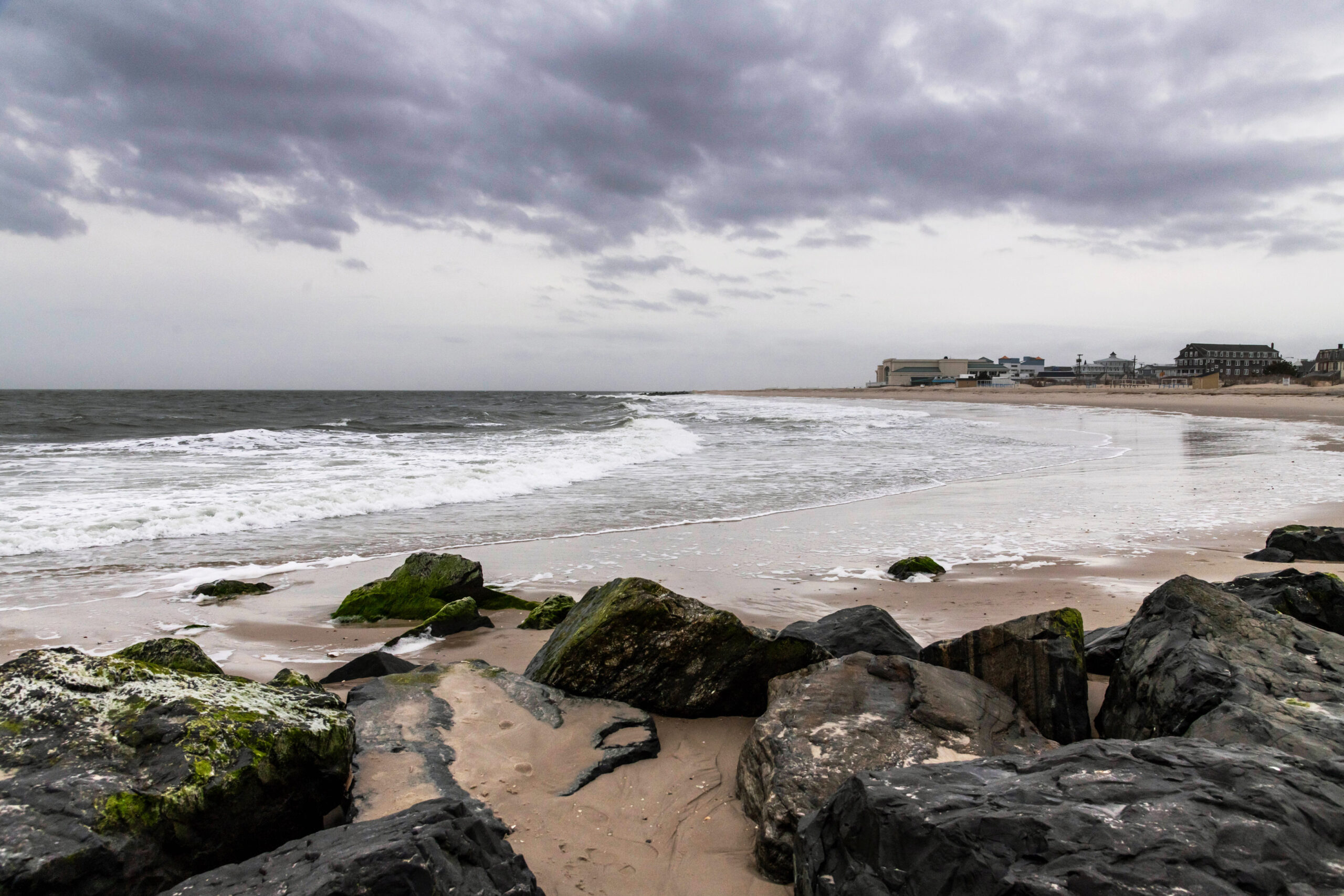 A wide view of a stormy cloudy day at the beach. Rocks are in the foreground, and Cape May Convention Hall is in the distance. The ocean is gray and foamy, and there is a dark gray cloud in a light gray sky.