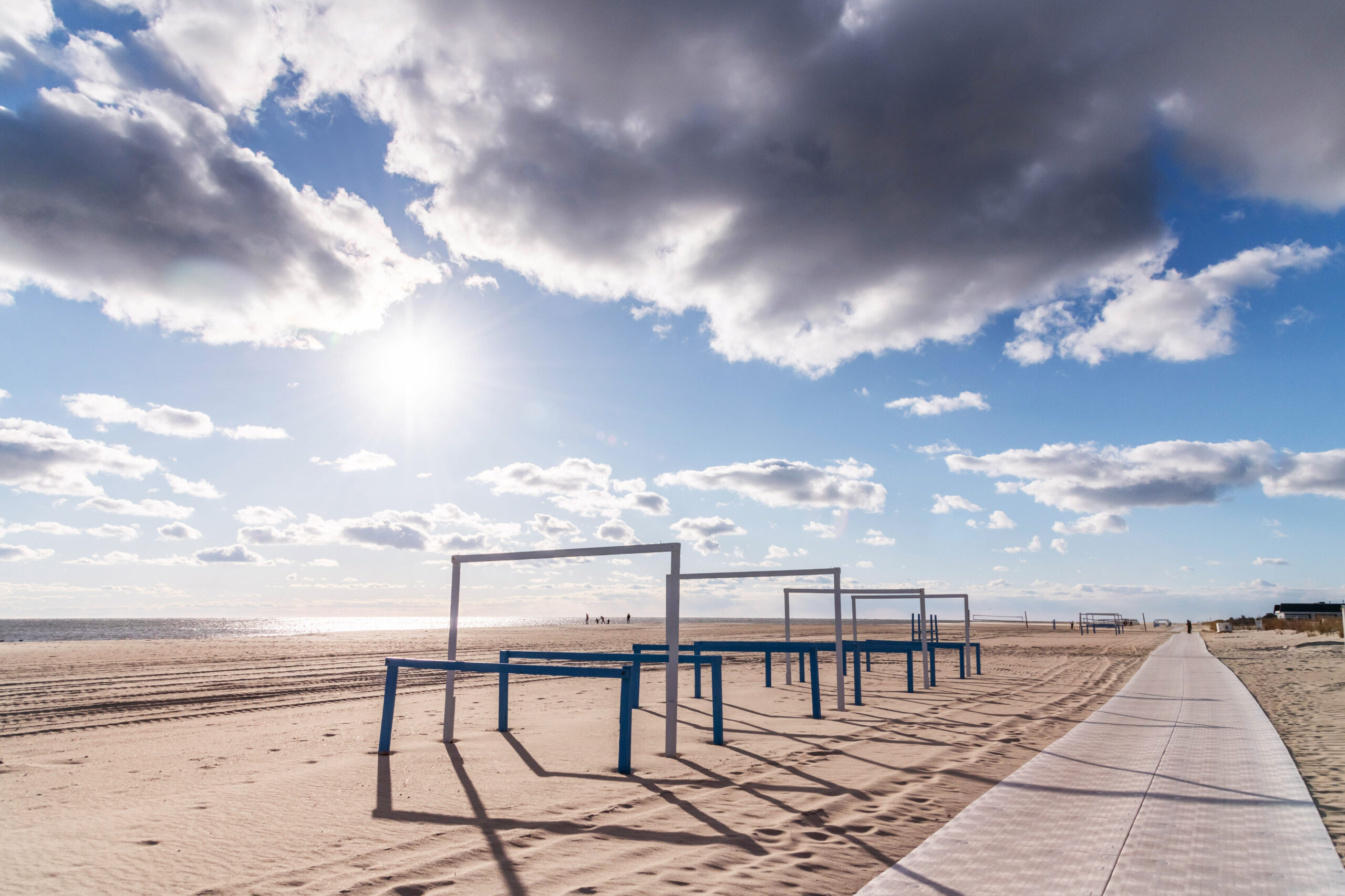 A wide view of the beach. There is a blue sky with puffy white clouds, and the sun is brightly shining. There are Steger's Beach Stands lined up in a row, casting shadows on the sand.