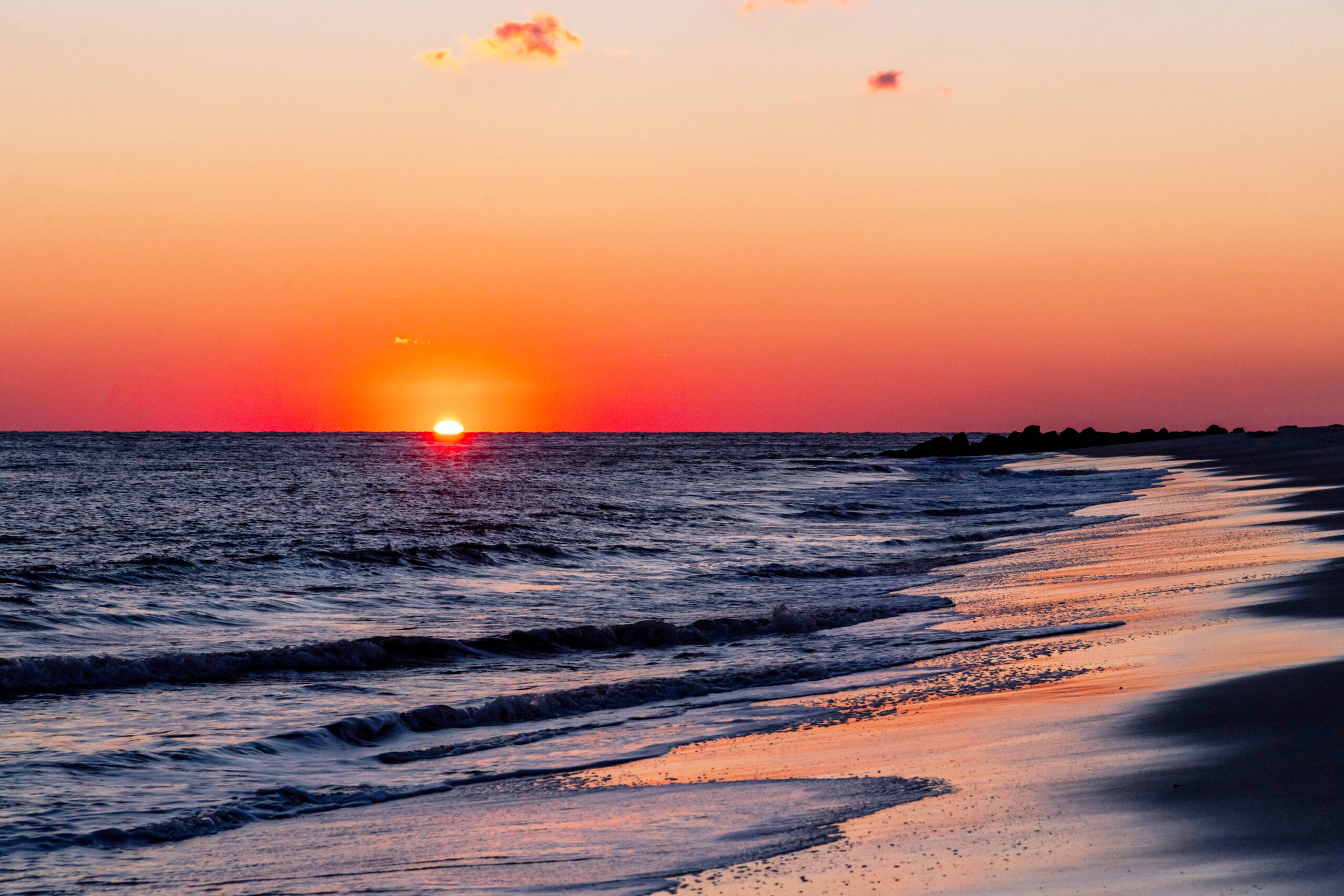 A wide view of the sun setting at the beach. The sun has half disappeared below the horizon line and ocean. The sky is clear and pink and orange. Waves are rushing into the shoreline.