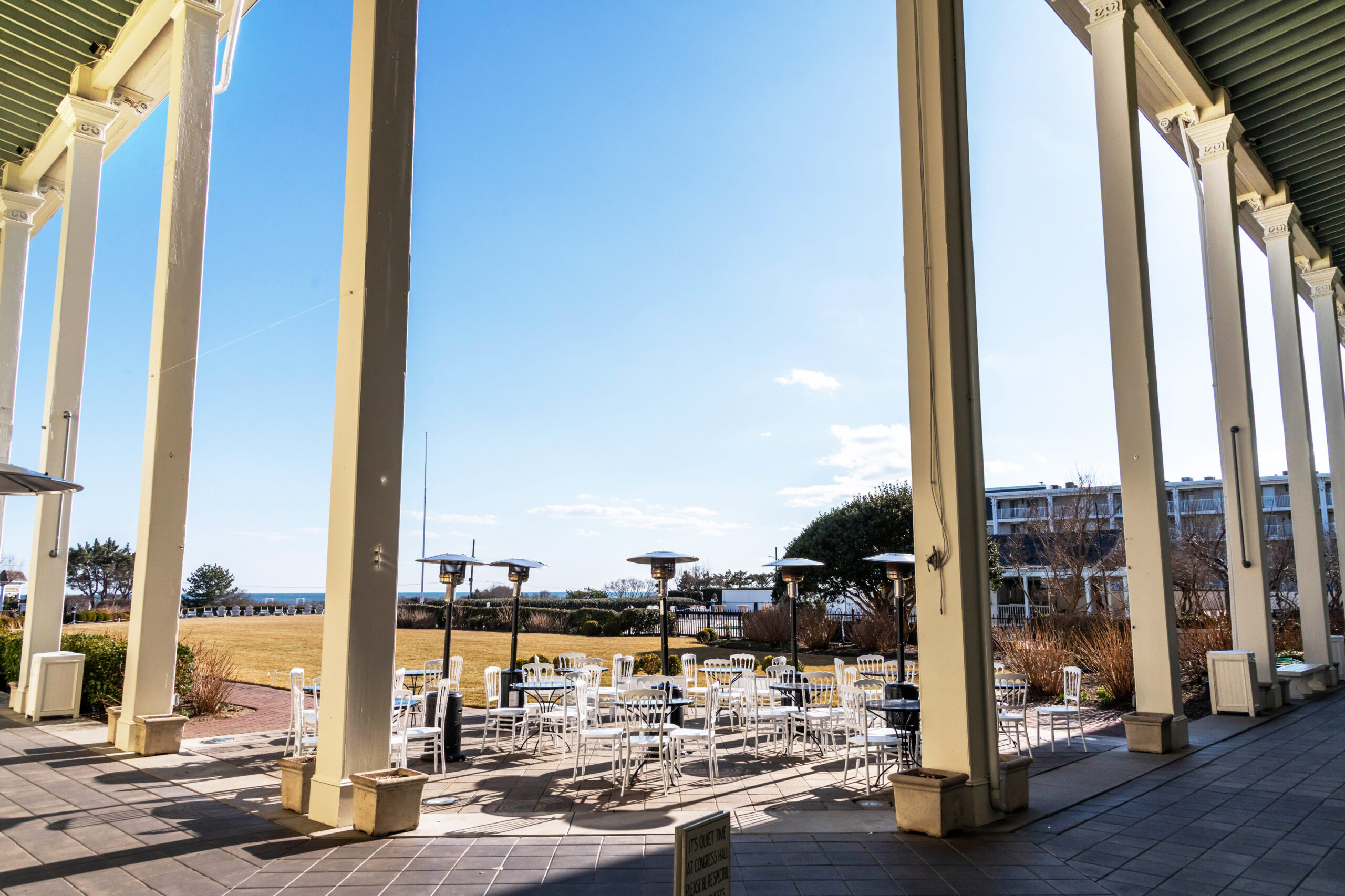 A wide view of the front lawn at Congress Hall. There are empty tables and chairs framed by columns. The sky is clear blue.