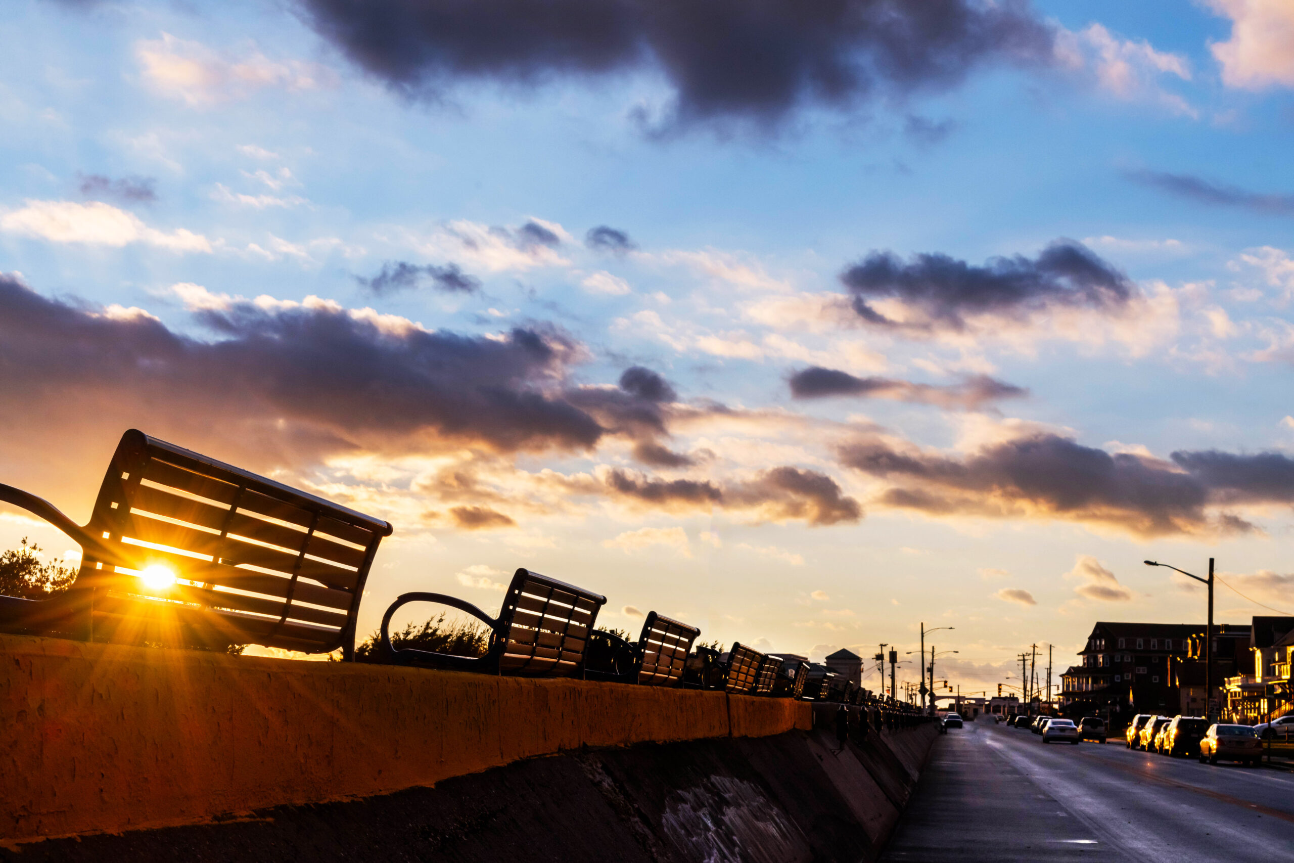 A wide view of the promenade and Beach Ave. The setting sun is shining behind a bench on the promenade. There are a few dark purple, gold, and white puffy clouds in the sky.